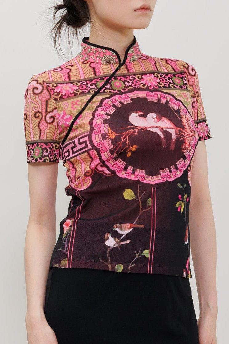 A model wearing a short-sleeved black midi skirt paired with a pink and black patterned top, styled with black pointed-toe heels against a light gray studio background.
