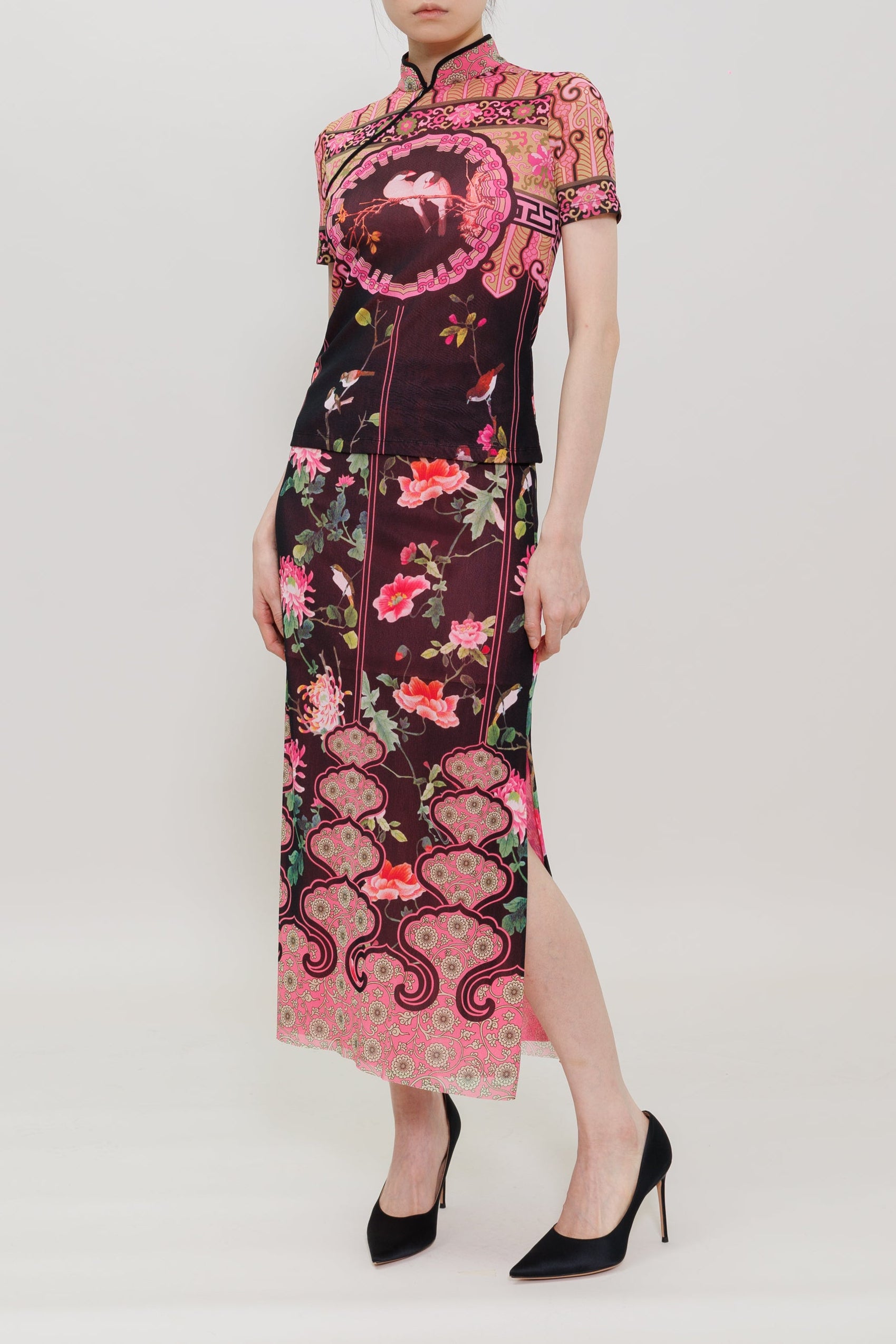 A model wears a short-sleeved, high-neck cheongsam-style dress with a dark plum base and vibrant pink floral and cloud motifs, paired with black high heels against a light gray studio background.