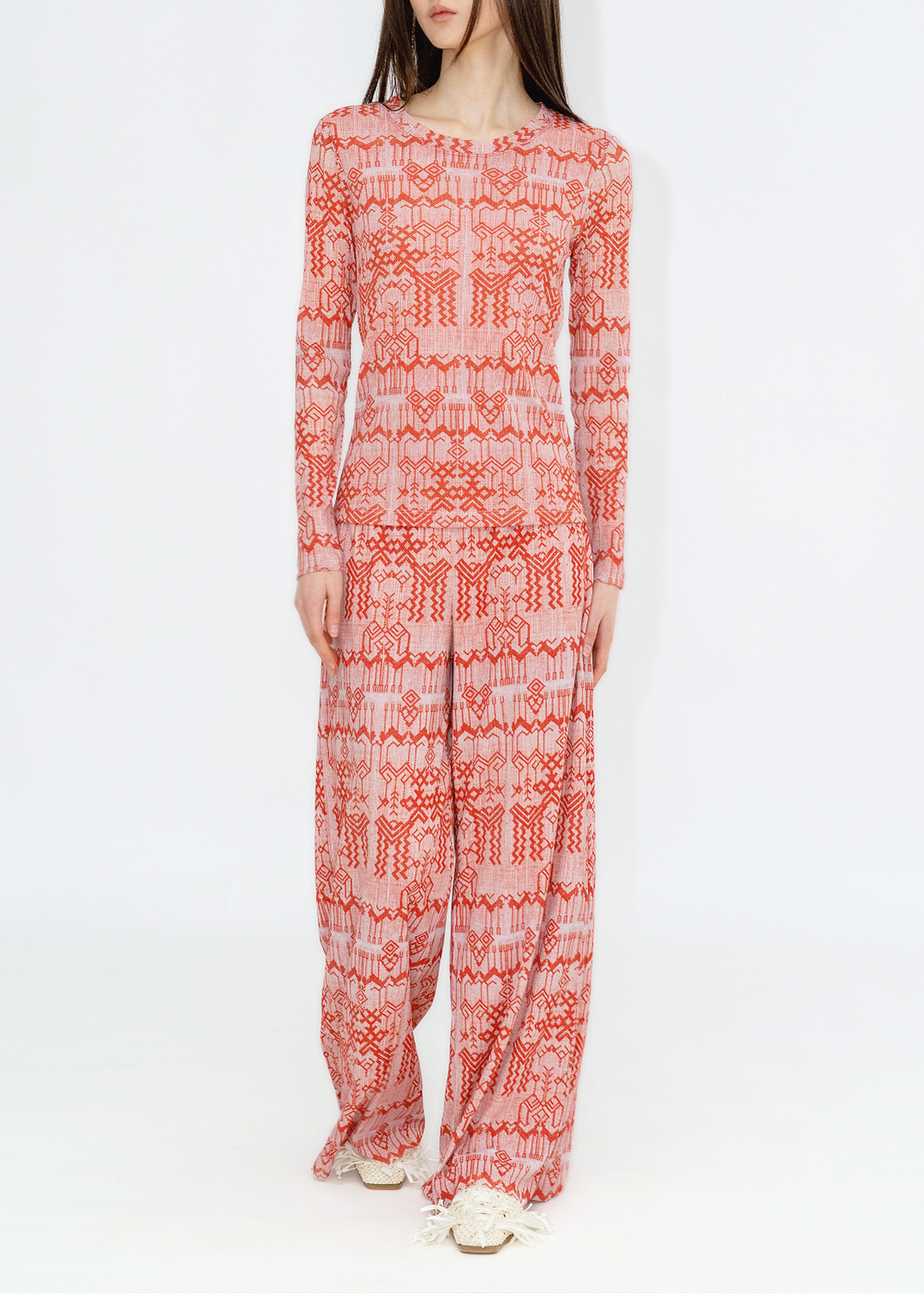 A woman modeling a long-sleeve, wide-leg jumpsuit with a red and white geometric pattern, photographed against a plain white studio background.
