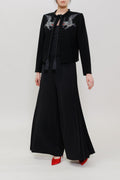 A model stands against a light gray studio background wearing a black outfit consisting of a cropped jacket with gray dragon embroidery on the shoulders, a black top, wide-leg black pants, and red pointed shoes.