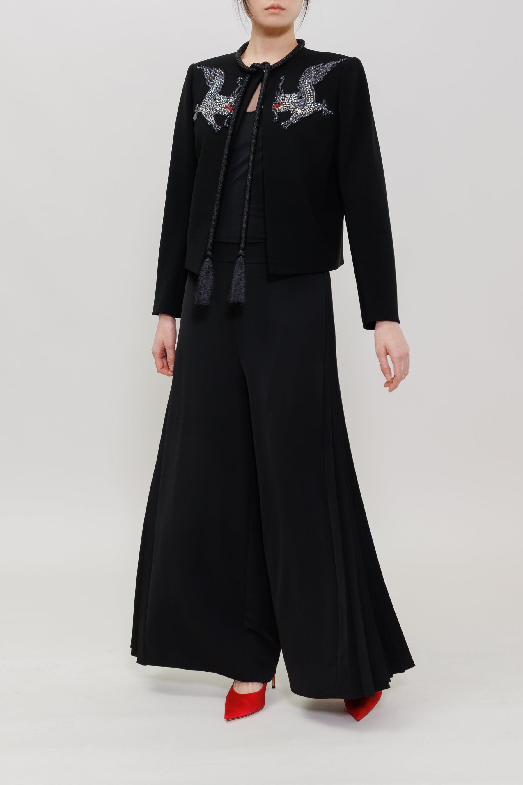 A model stands against a light gray studio background wearing a black outfit consisting of a cropped jacket with gray dragon embroidery on the shoulders, a black top, wide-leg black pants, and red pointed shoes.