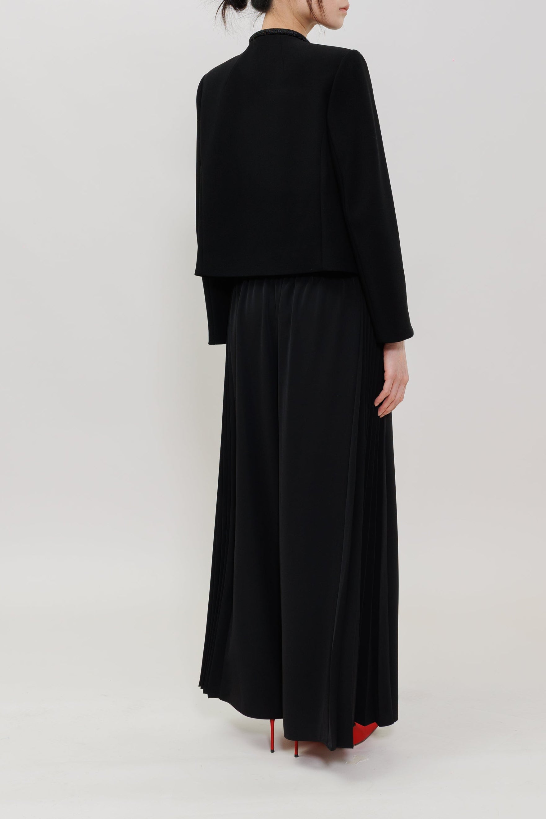 A model is shown from the back wearing a black long-sleeve cropped jacket and a long black pleated skirt, paired with red high heels, against a plain white studio background.