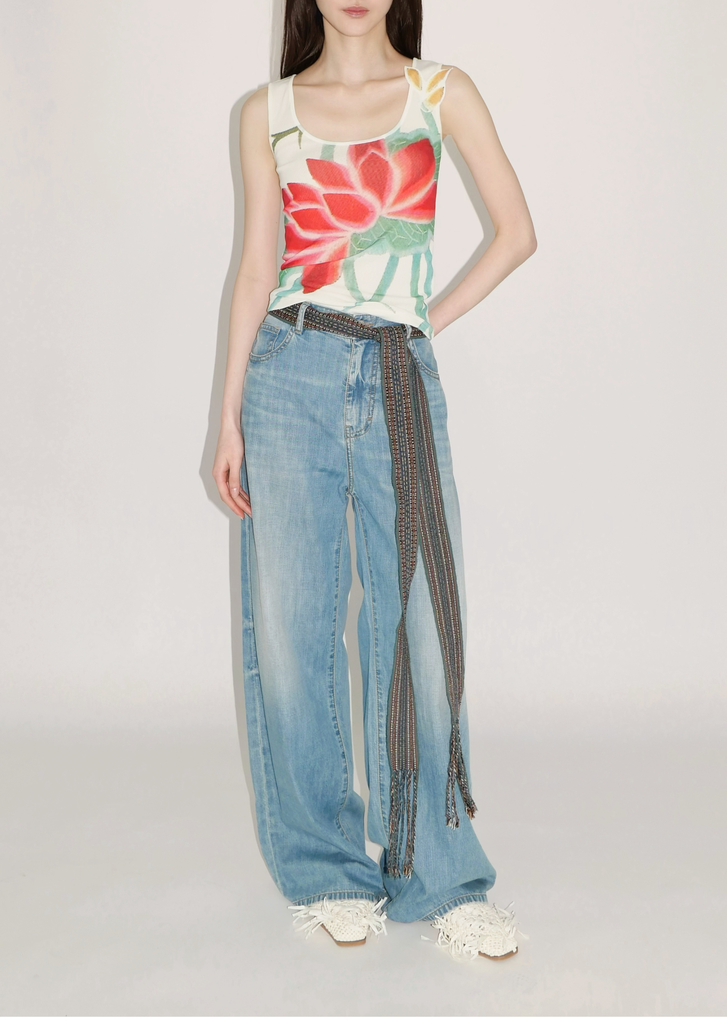 A model stands against a light gray studio background wearing a sleeveless tank top with a large red and green floral print on a cream base, paired with wide-leg light blue jeans featuring a dark patterned tie detail at the waist and white sneakers.