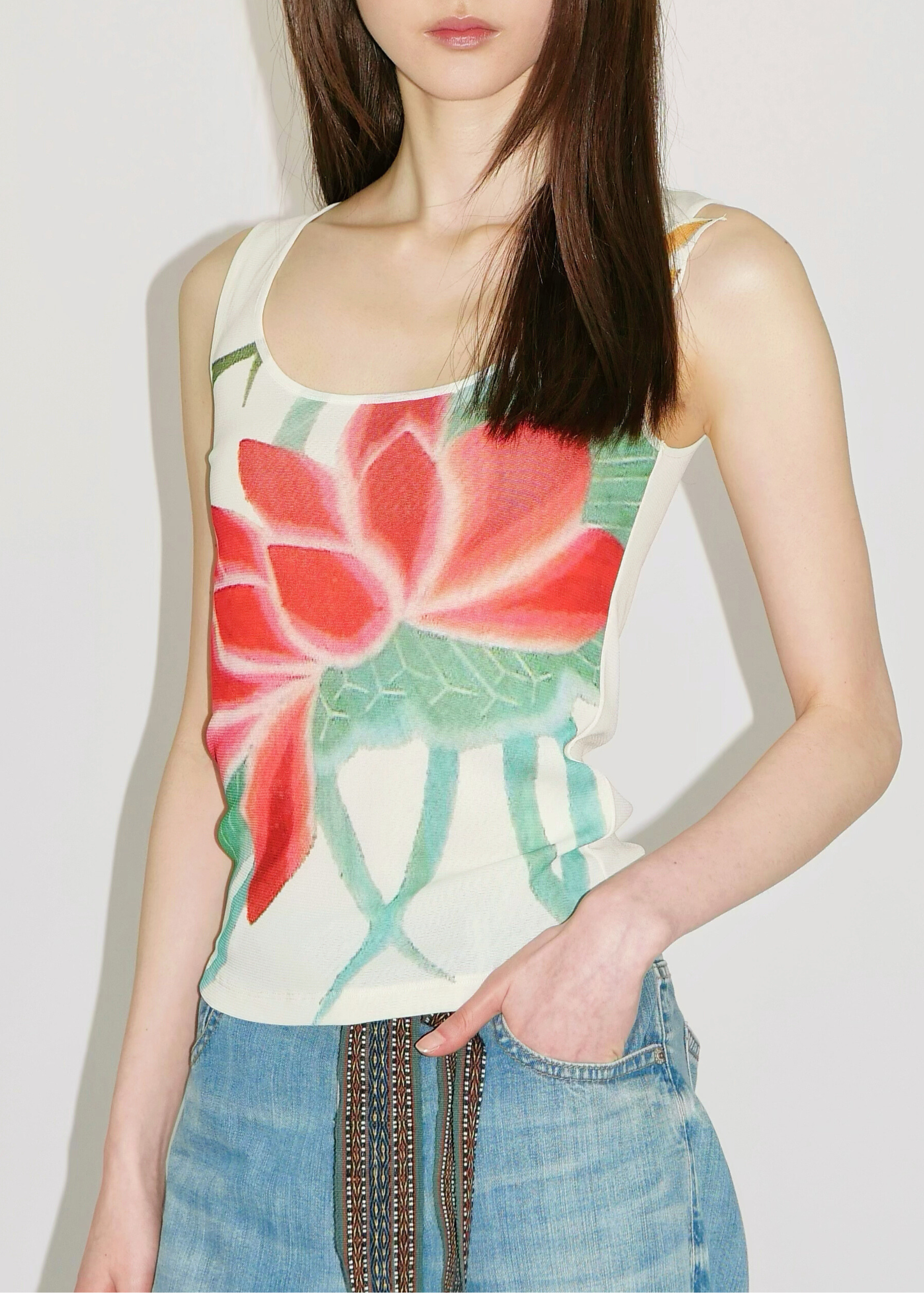 A woman wearing a sleeveless white tank top with a large red and green lotus flower print, paired with blue jeans featuring a decorative brown and gold zipper detail.