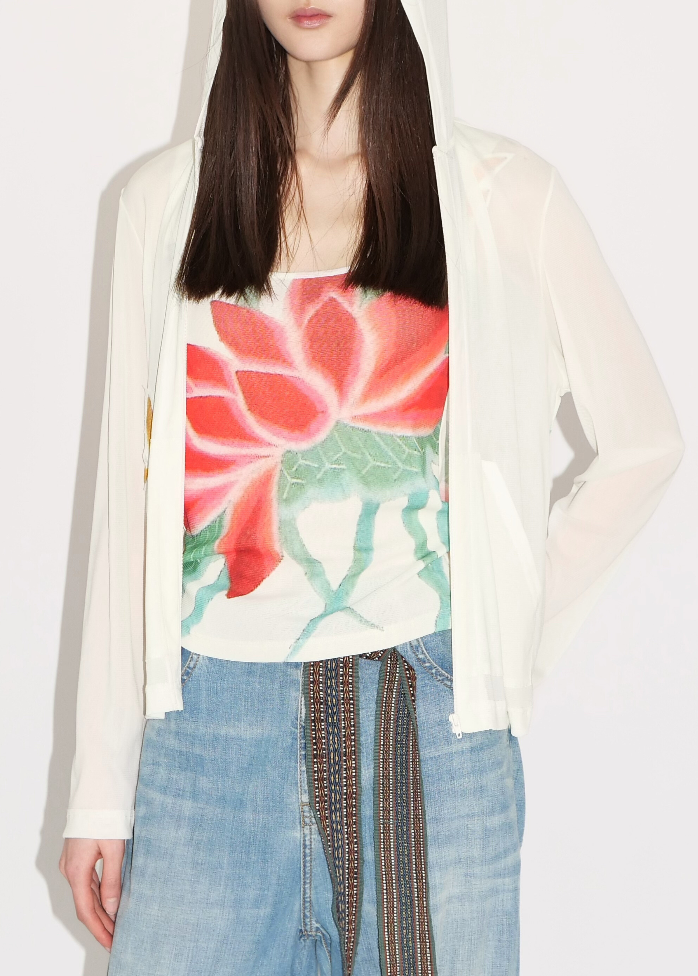 A model wearing a white hooded cardigan over a white tank top with a large red and green lotus flower print, paired with light blue jeans.