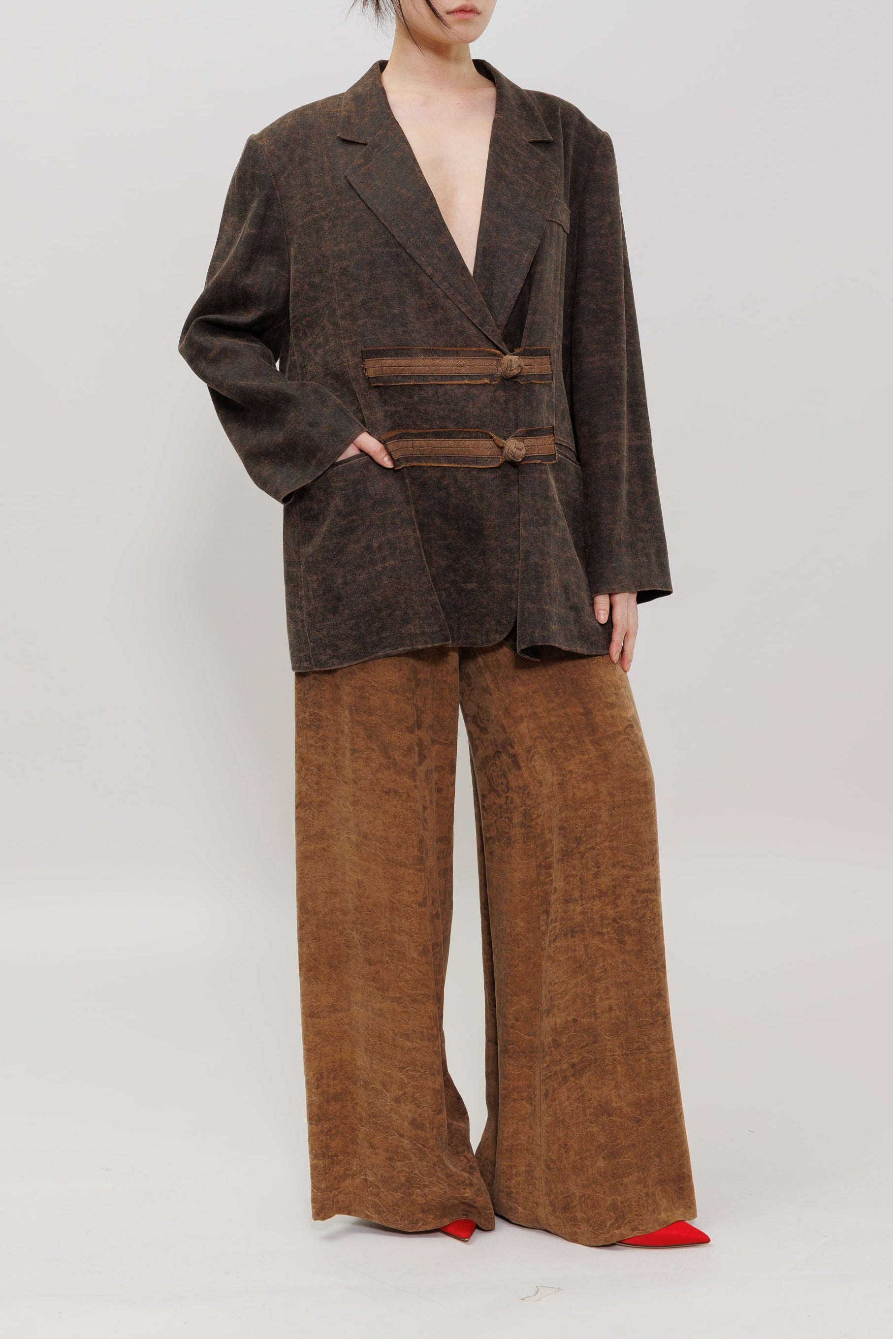A model stands against a light gray studio background wearing a dark brown textured blazer with a wrap-style double-breasted design and matching wide-leg brown trousers.