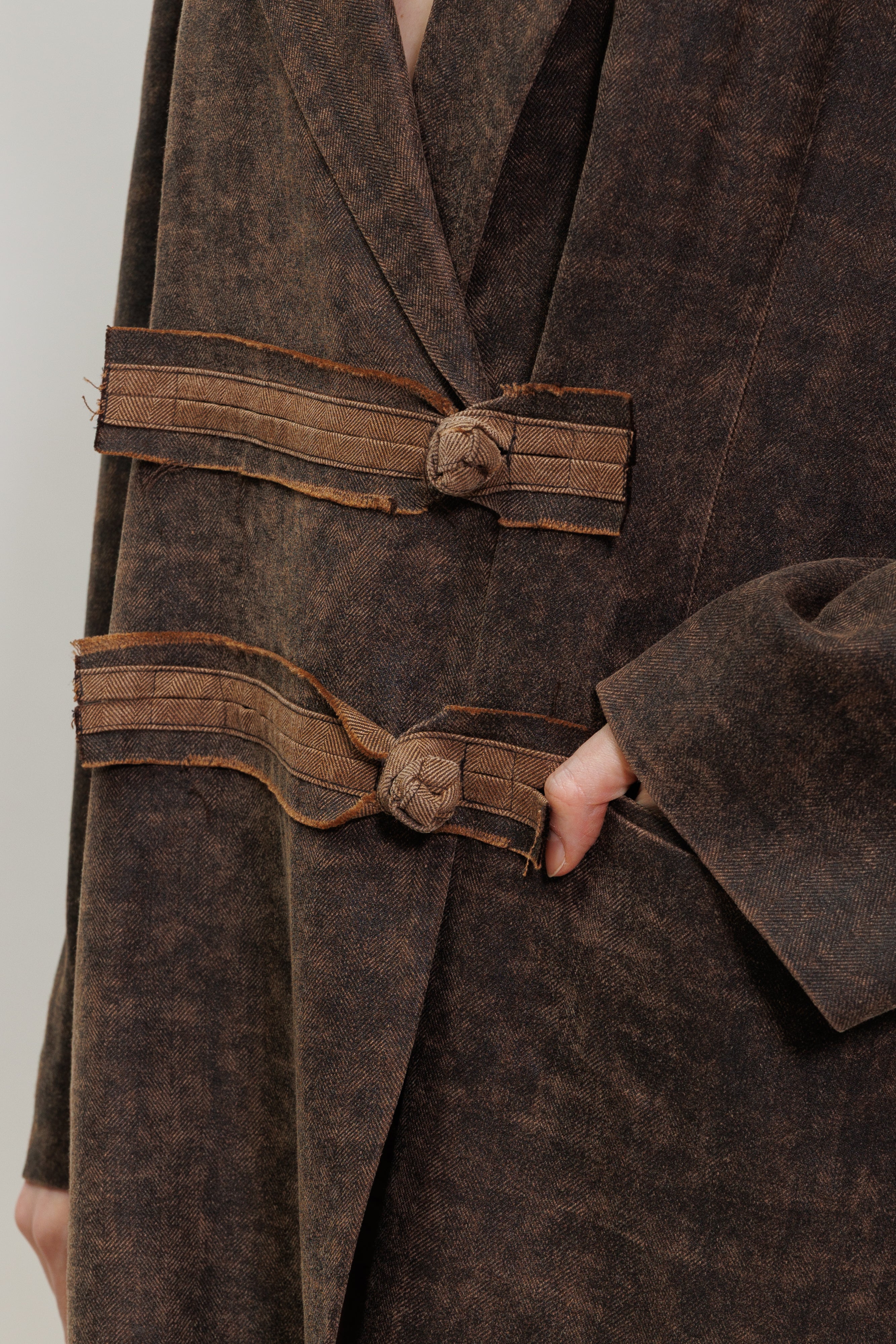 Close-up of a dark brown textured coat with wide decorative straps and knotted toggle details, worn by a model whose hand is visible.
