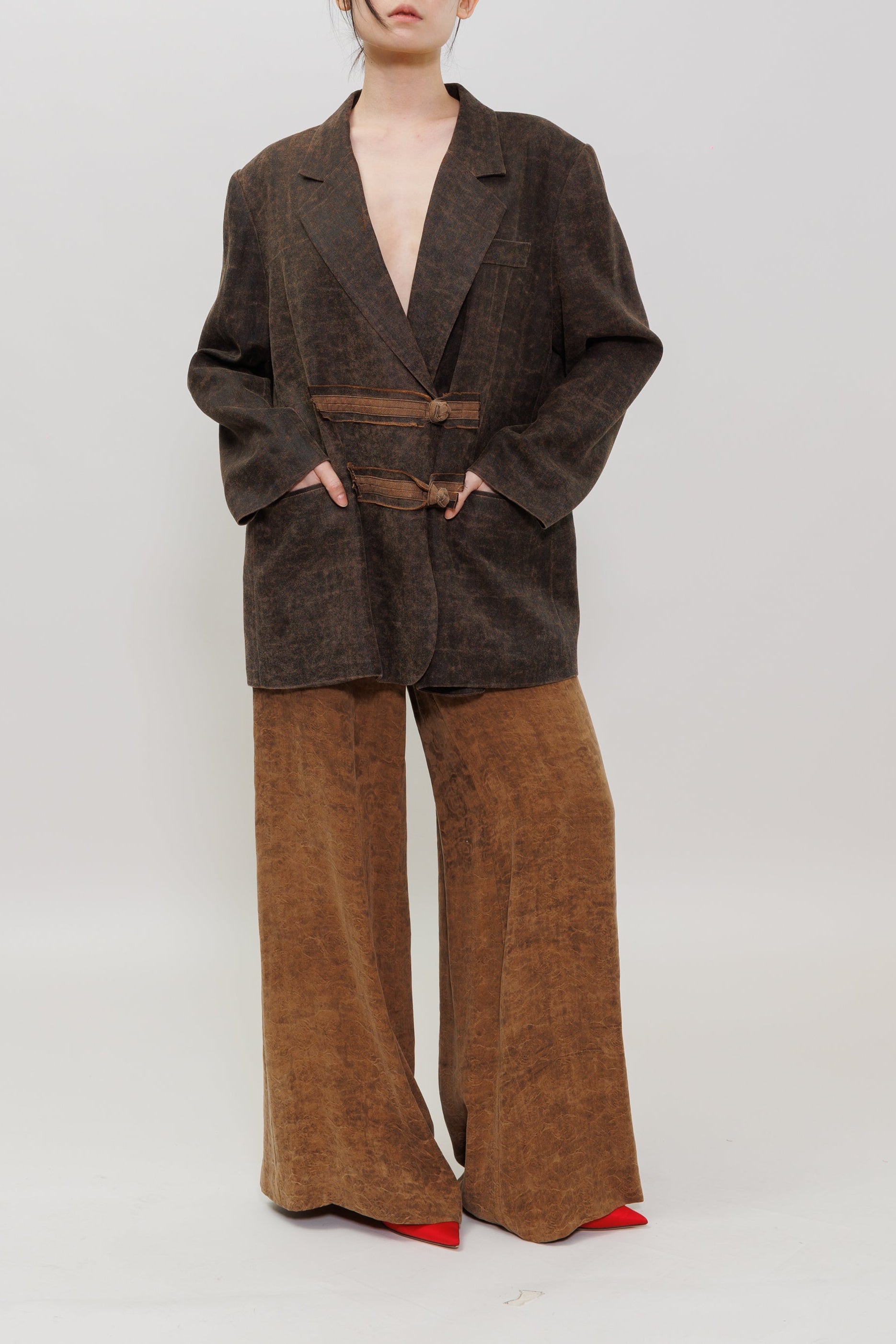 A model stands against a light gray studio background wearing a dark brown textured blazer, wide-leg brown pants, and red pointed shoes.