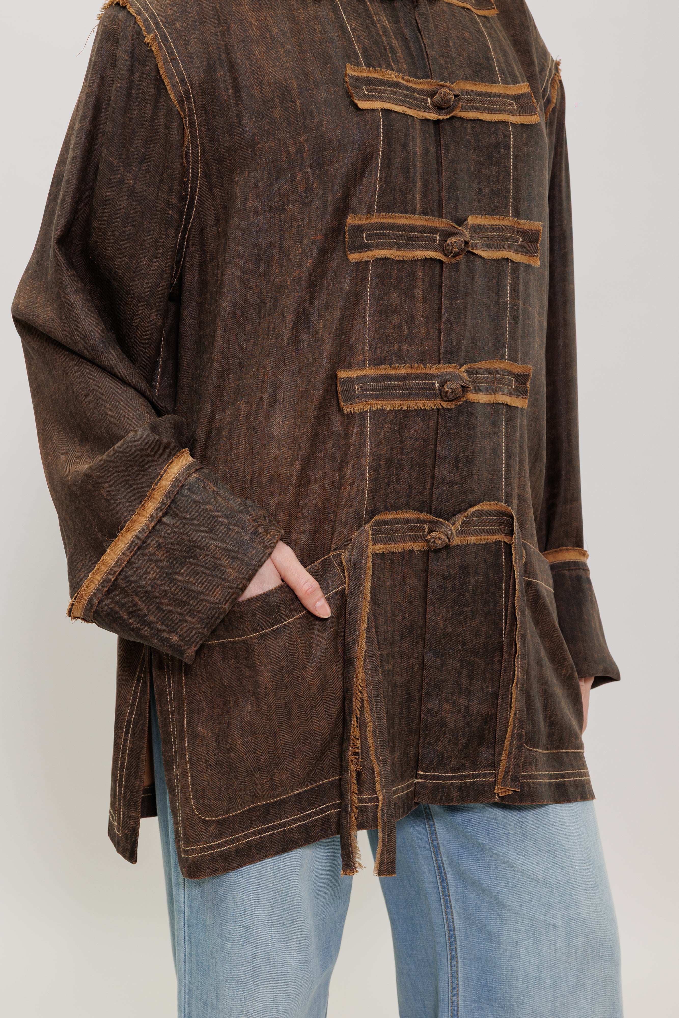 A dark brown, long-sleeved tunic top with a distressed, mineral-washed texture, featuring decorative button closures and contrast stitching, styled with light blue jeans.