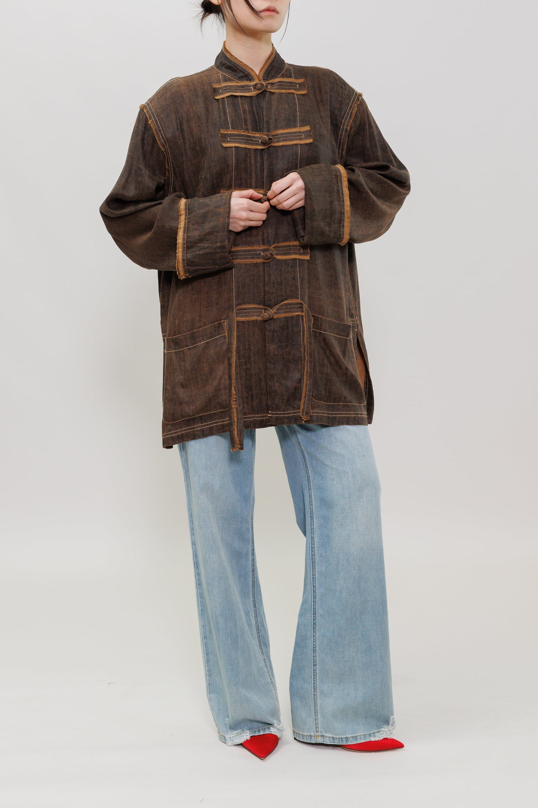 A model stands against a light gray studio background wearing a dark brown corduroy jacket with gold-colored contrast stitching and traditional Chinese-style frog closures, paired with light blue wide-leg jeans and red pointed shoes.