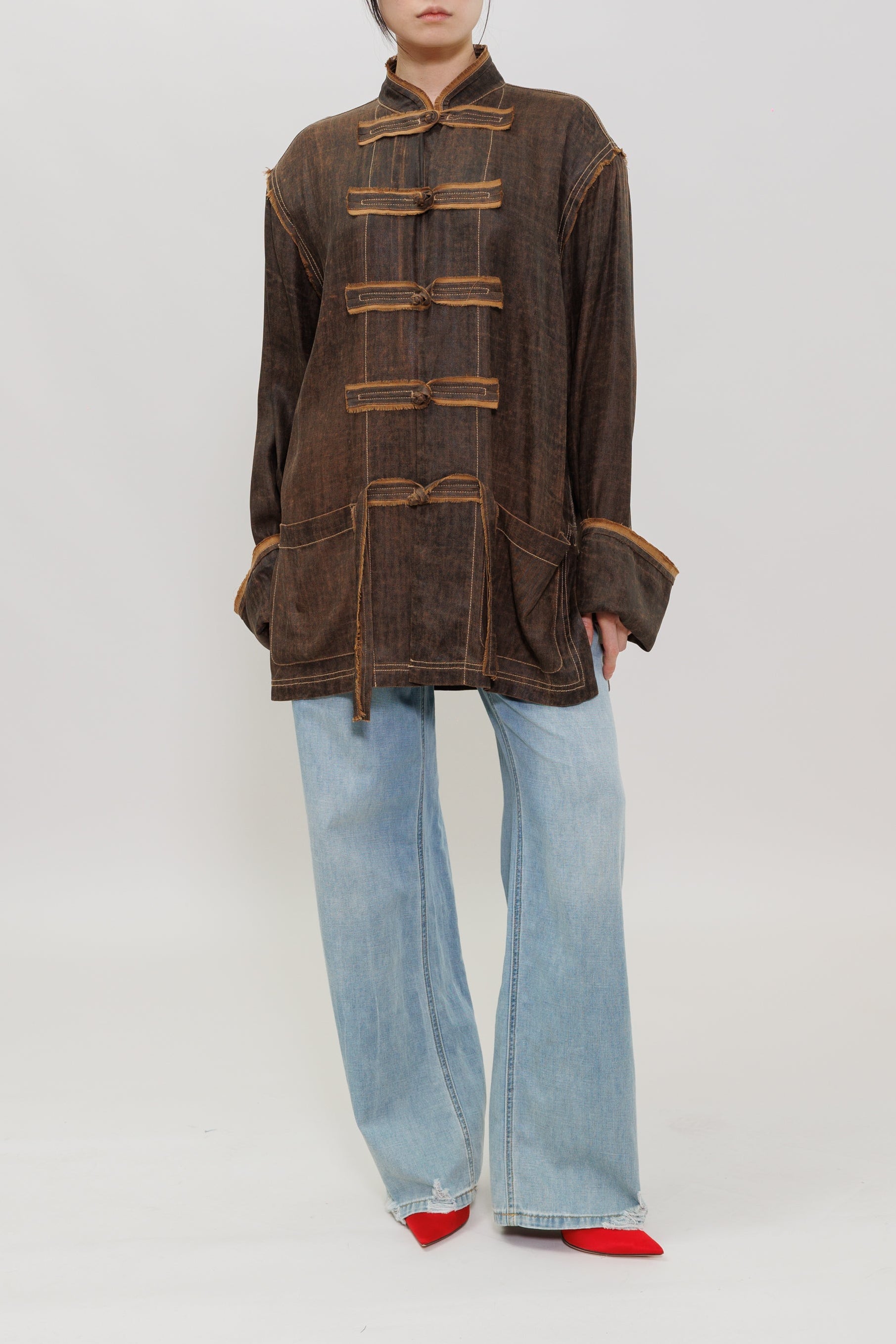 A model stands against a light gray studio background wearing a brown cropped jacket with tan contrast stitching and traditional frog closures, paired with light blue wide-leg jeans and red pointed shoes.