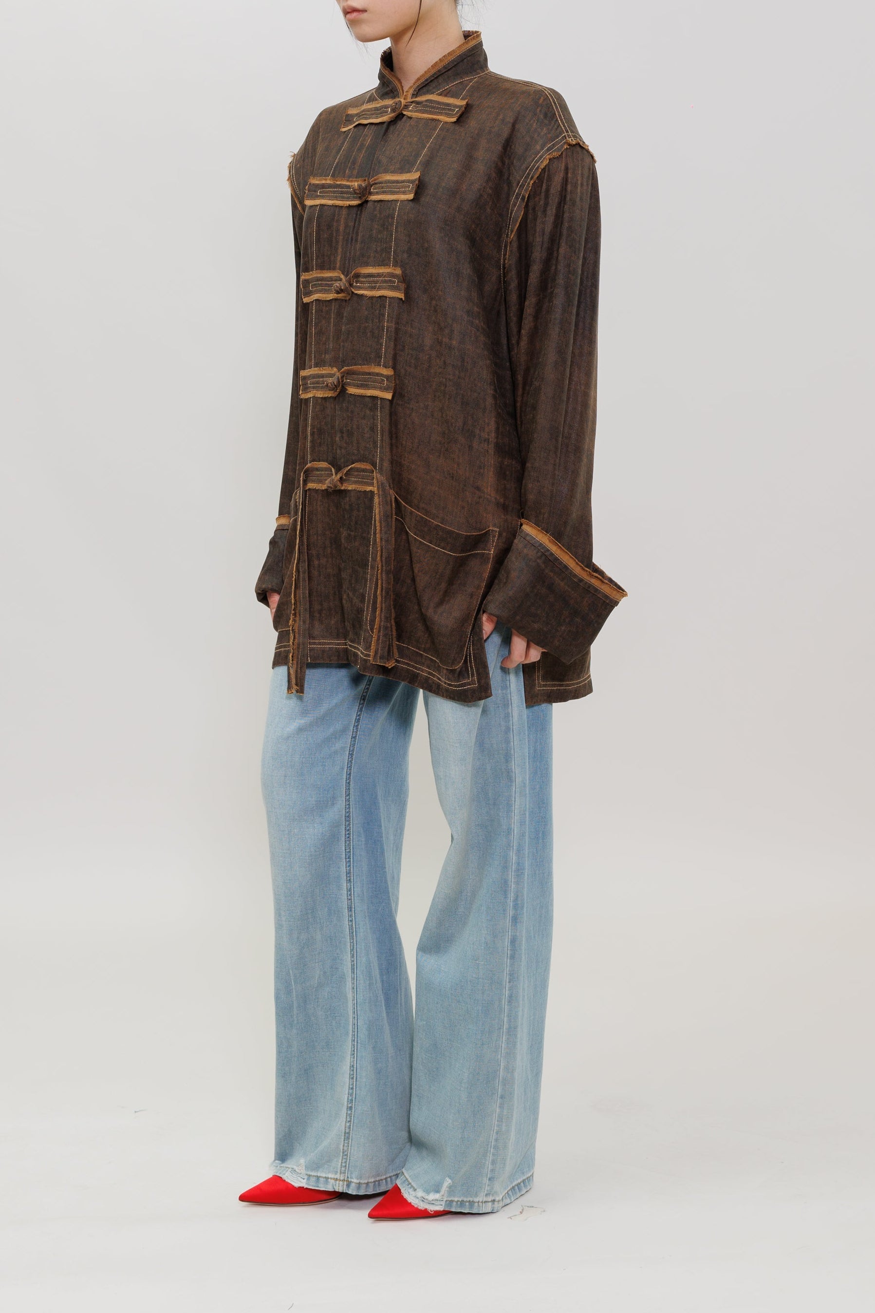 A model stands in profile wearing a dark brown textured jacket with gold-toned decorative trim and buttons, paired with light blue wide-leg jeans and red shoes, against a plain light gray studio background.