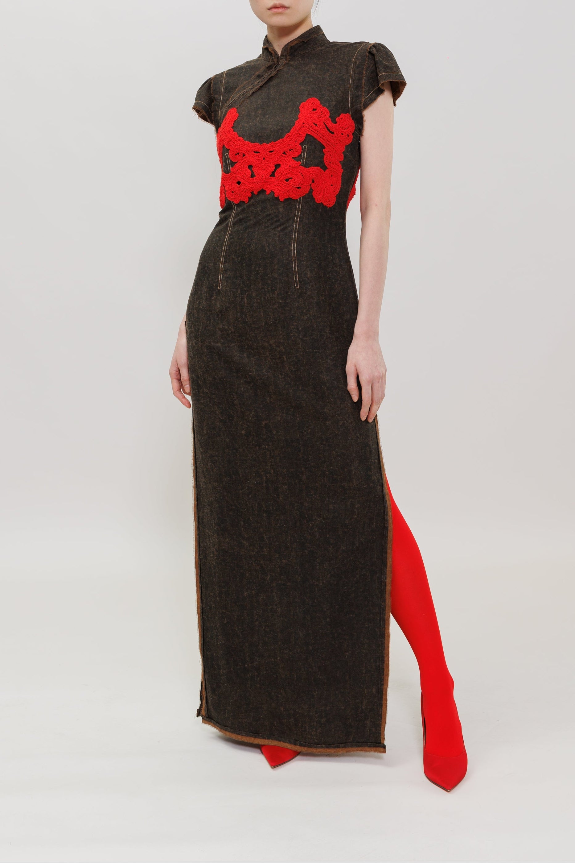 A model stands against a plain light gray studio background wearing a dark brown textured cheongsam-style dress with a bright red embroidered chest motif and a red slit at the hem.