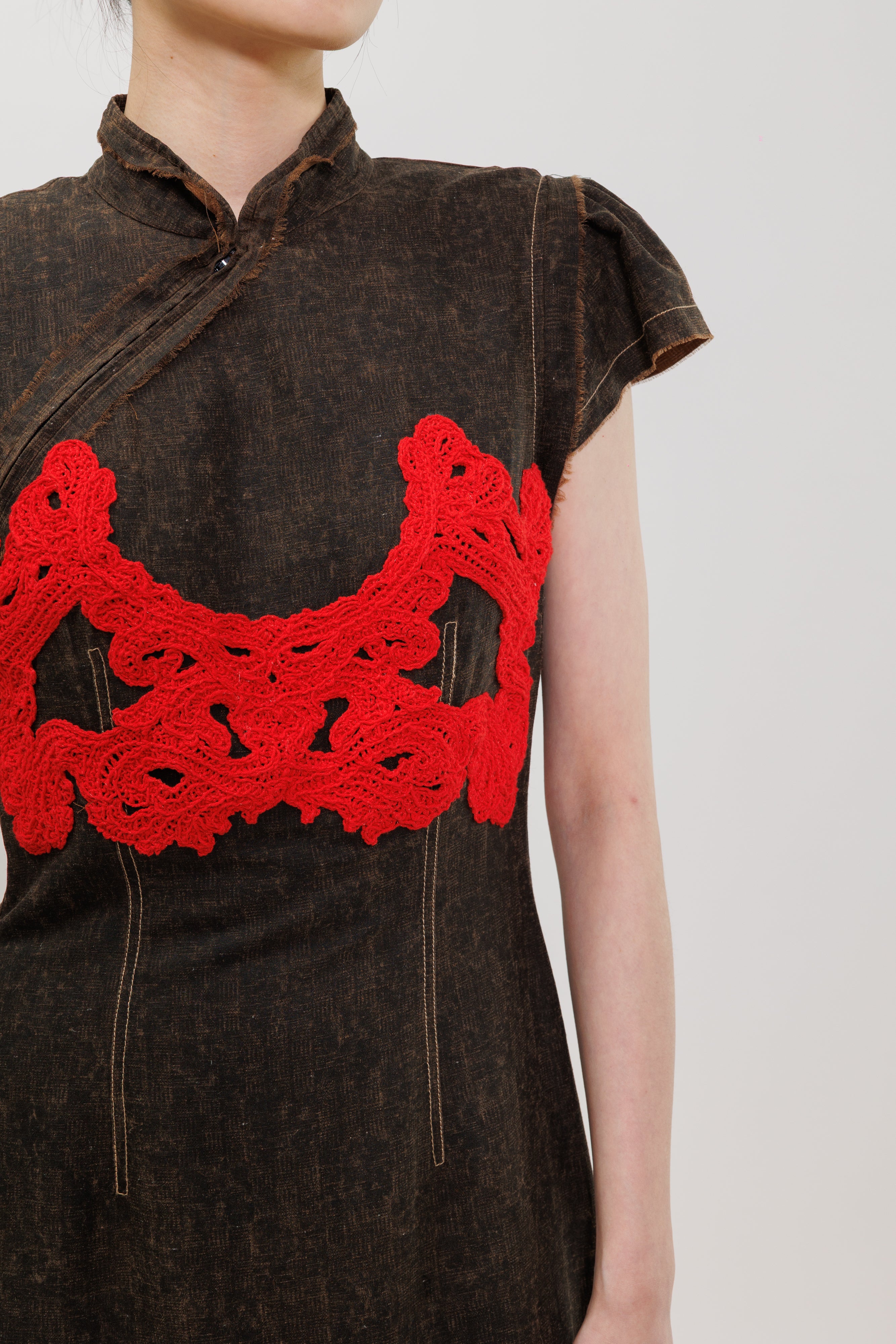Close-up of a person wearing a dark brown textured dress with a bright red crocheted chest panel and a high collar, against a light gray background.