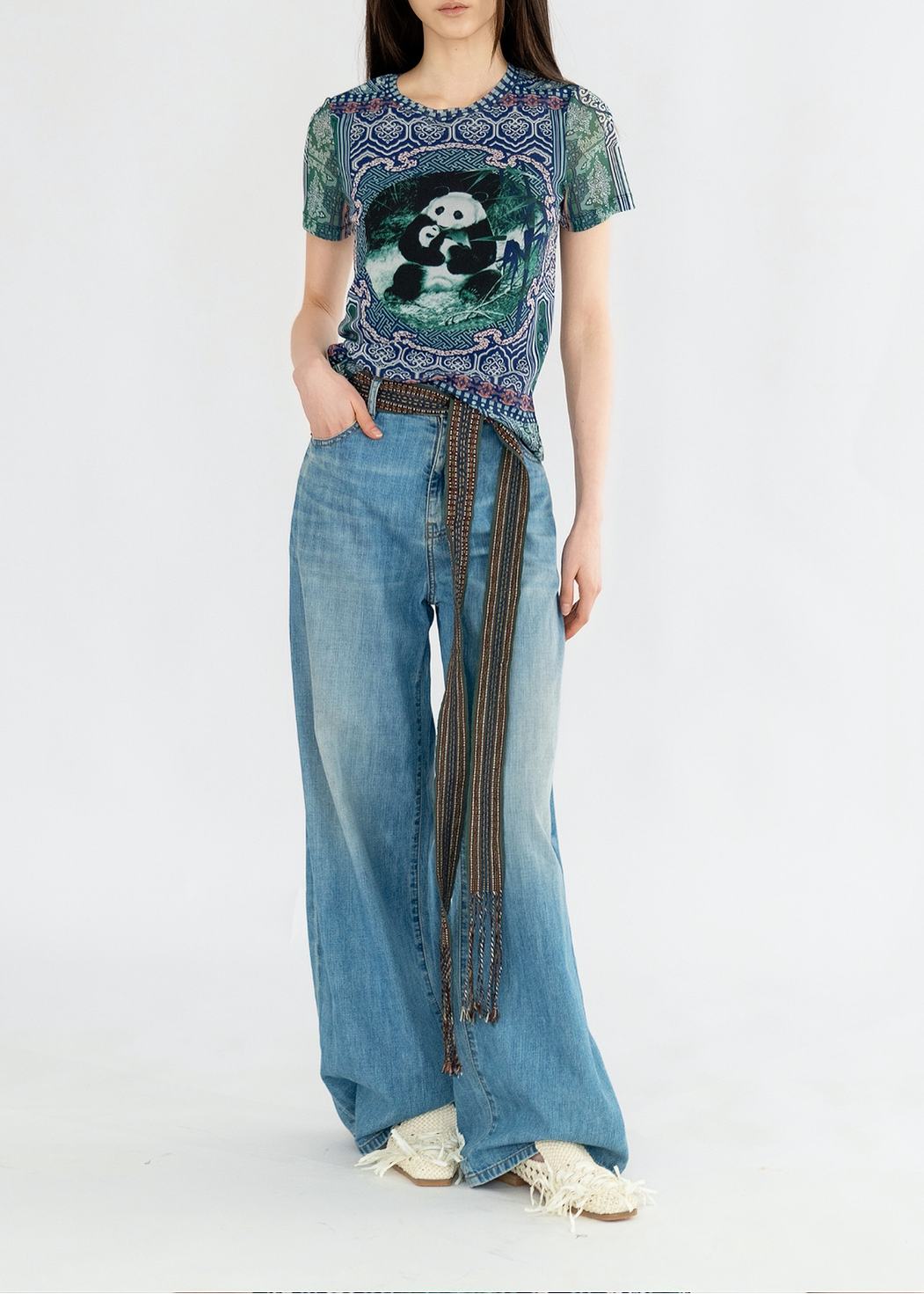 A woman is standing, wearing a multicolored netting top with a panda graphic and paired with blue denim jeans.