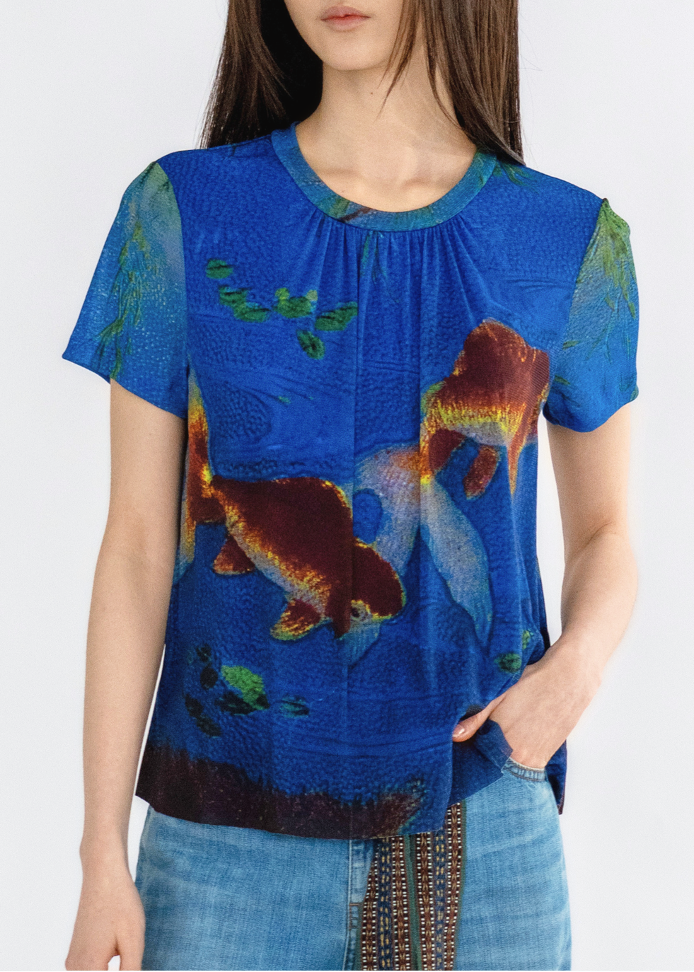 A woman wearing a blue top with a ruffled neckline and a pattern of goldfish and aquatic plants.