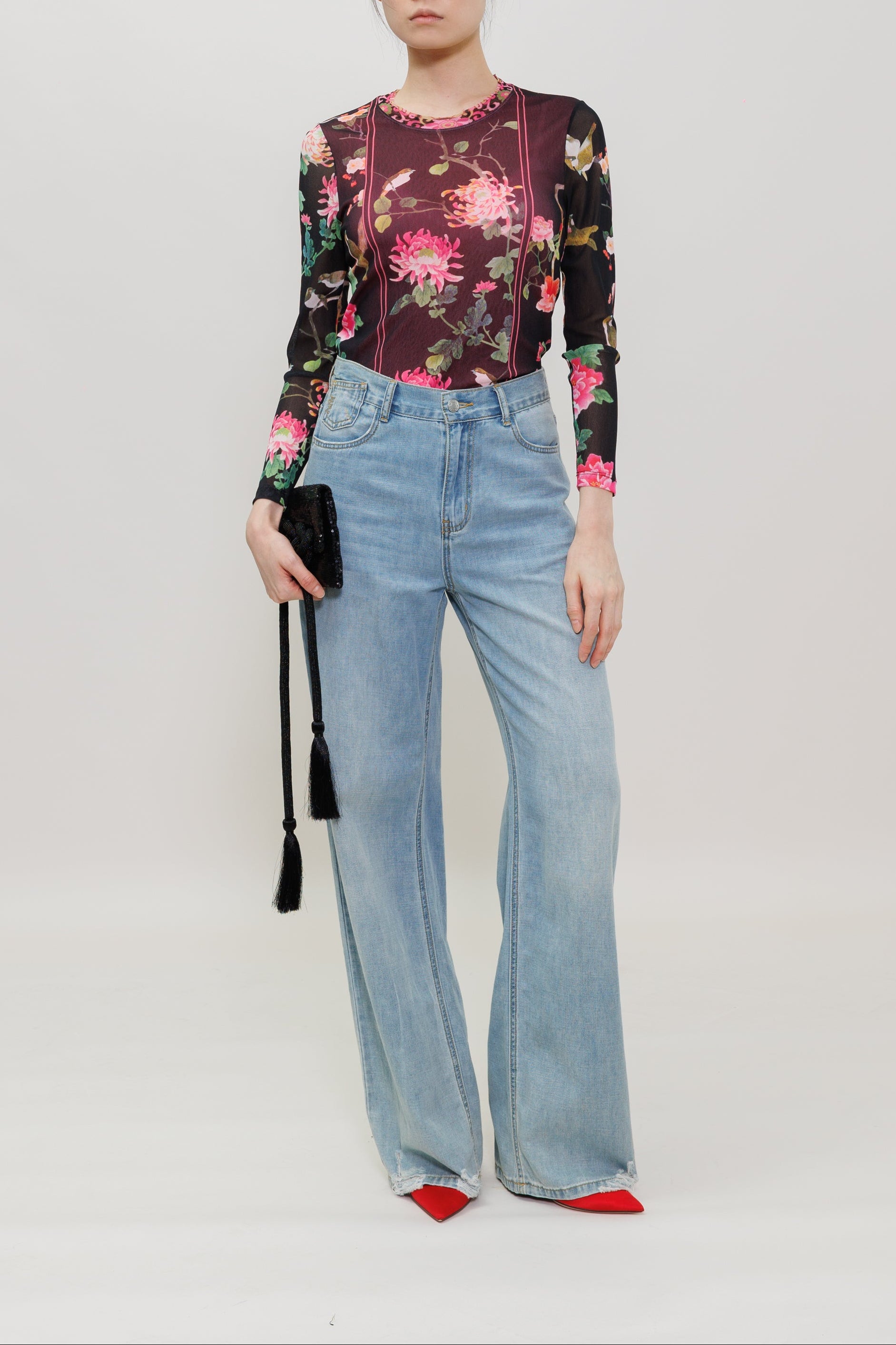 A model wearing a dark floral long-sleeve top, light-wash wide-leg jeans, and red pointed shoes, holding a black tassel clutch against a plain light gray studio background.