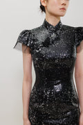 A close-up of a model wearing a black sequined dress with sheer flutter sleeves, a high collar, and a black fabric flower detail on the chest, set against a plain light gray background.