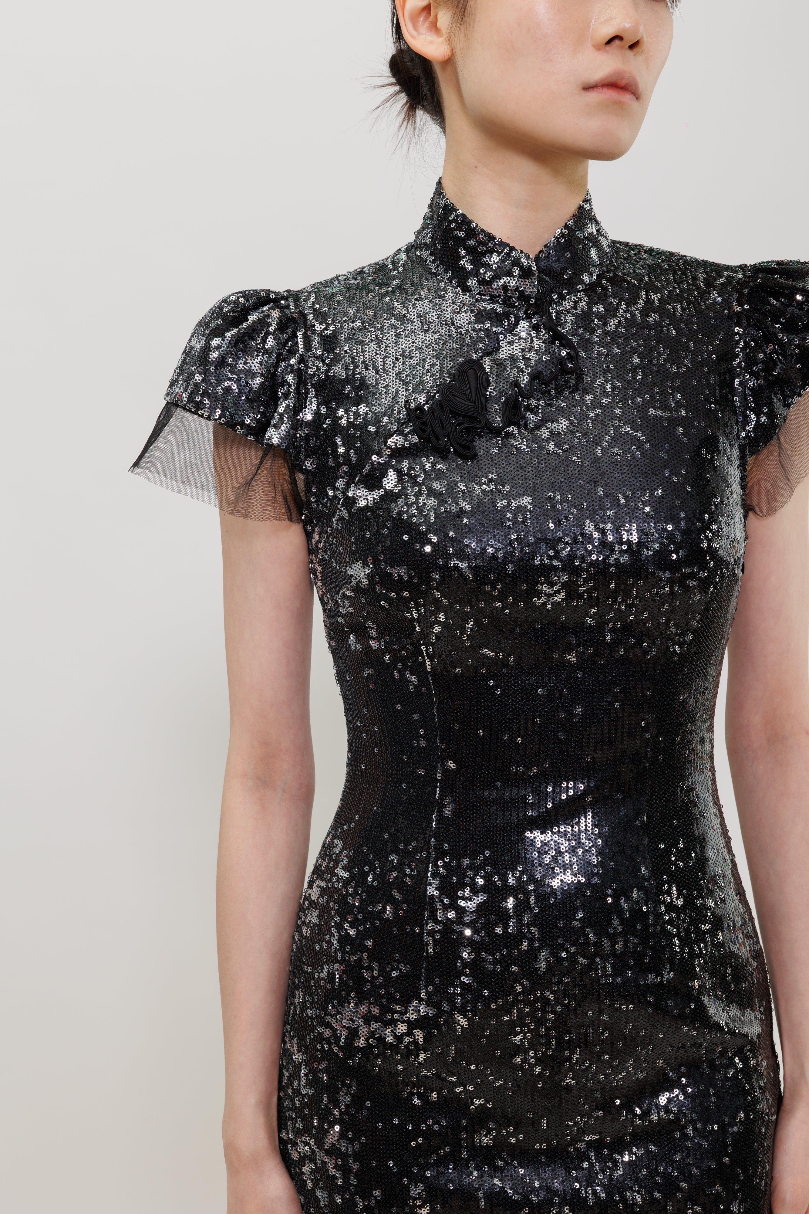 A close-up of a model wearing a black sequined dress with sheer flutter sleeves, a high collar, and a black fabric flower detail on the chest, set against a plain light gray background.
