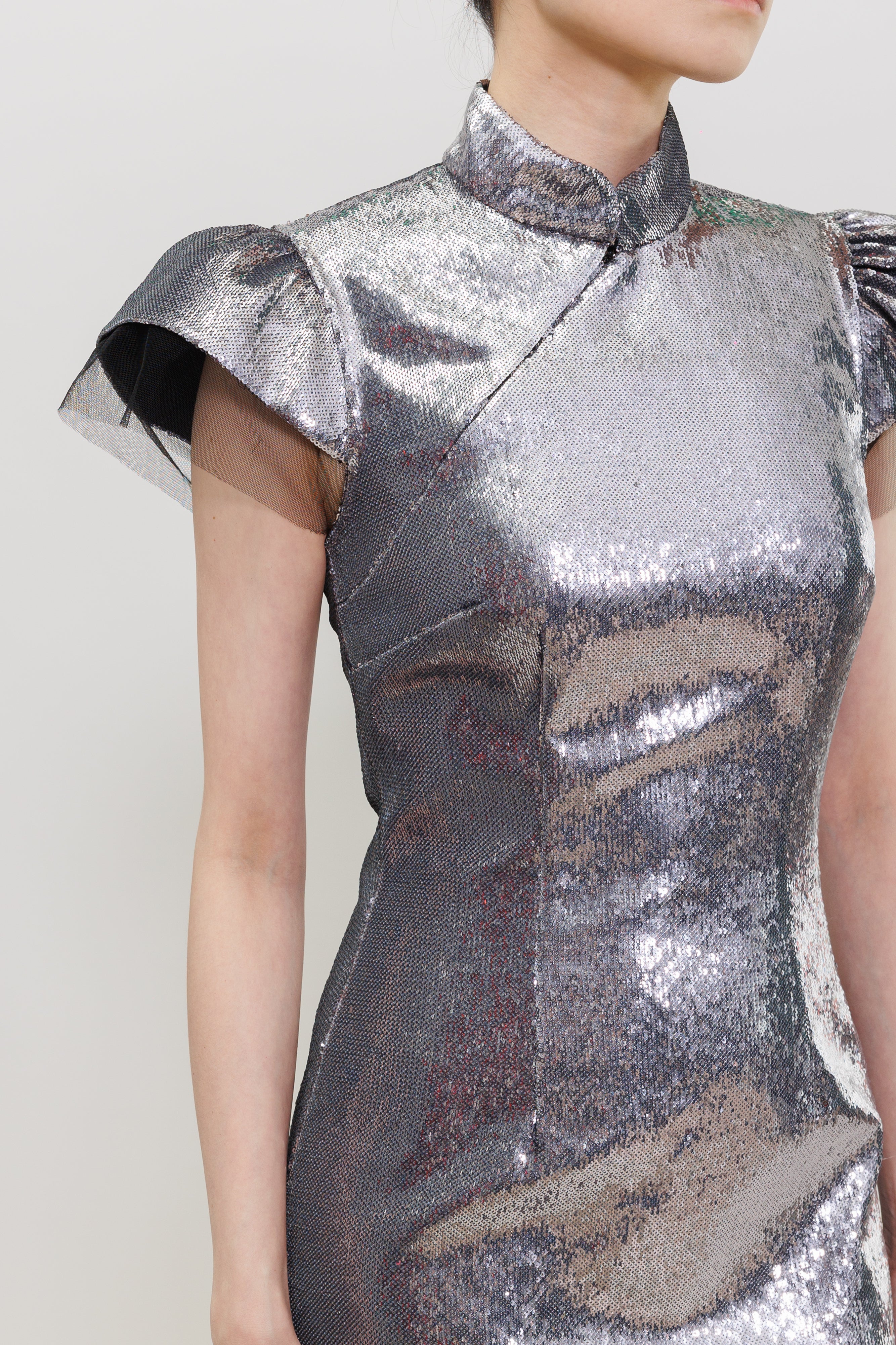 A close-up of a model wearing a metallic silver cheongsam-style dress with ruffled cap sleeves and a high collar, set against a plain light gray background.