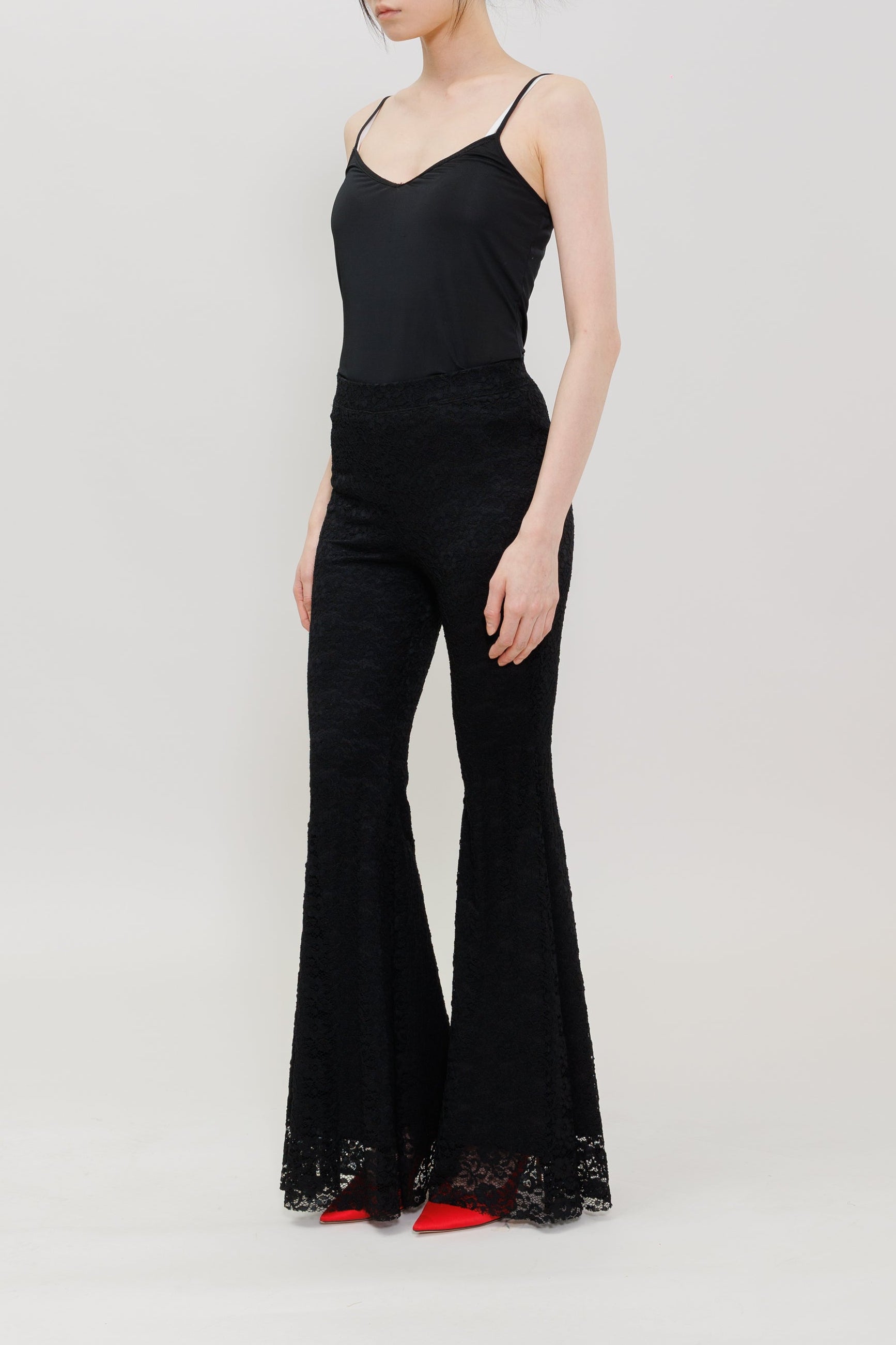 A woman modeling a black sleeveless camisole top paired with matching black flared pants featuring lace trim at the hems, standing against a plain light gray studio background.