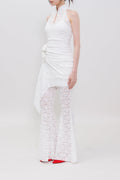 A model stands in profile wearing a white sleeveless halter dress with a ruched bodice, draped side detail, and matching white lace flared pants, against a plain light gray studio background.