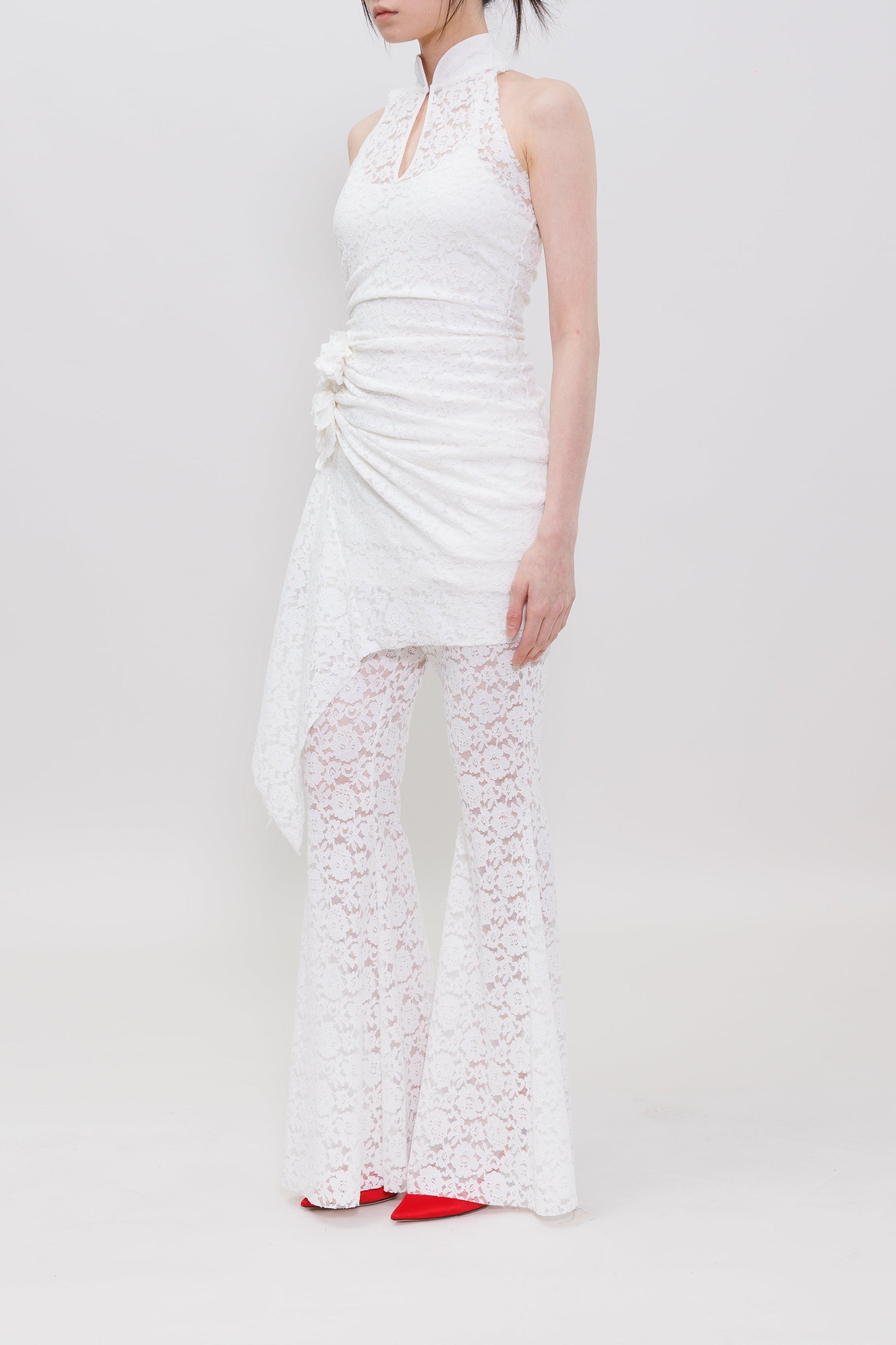 A model stands in profile wearing a white sleeveless halter dress with a ruched bodice, draped side detail, and matching white lace flared pants, against a plain light gray studio background.