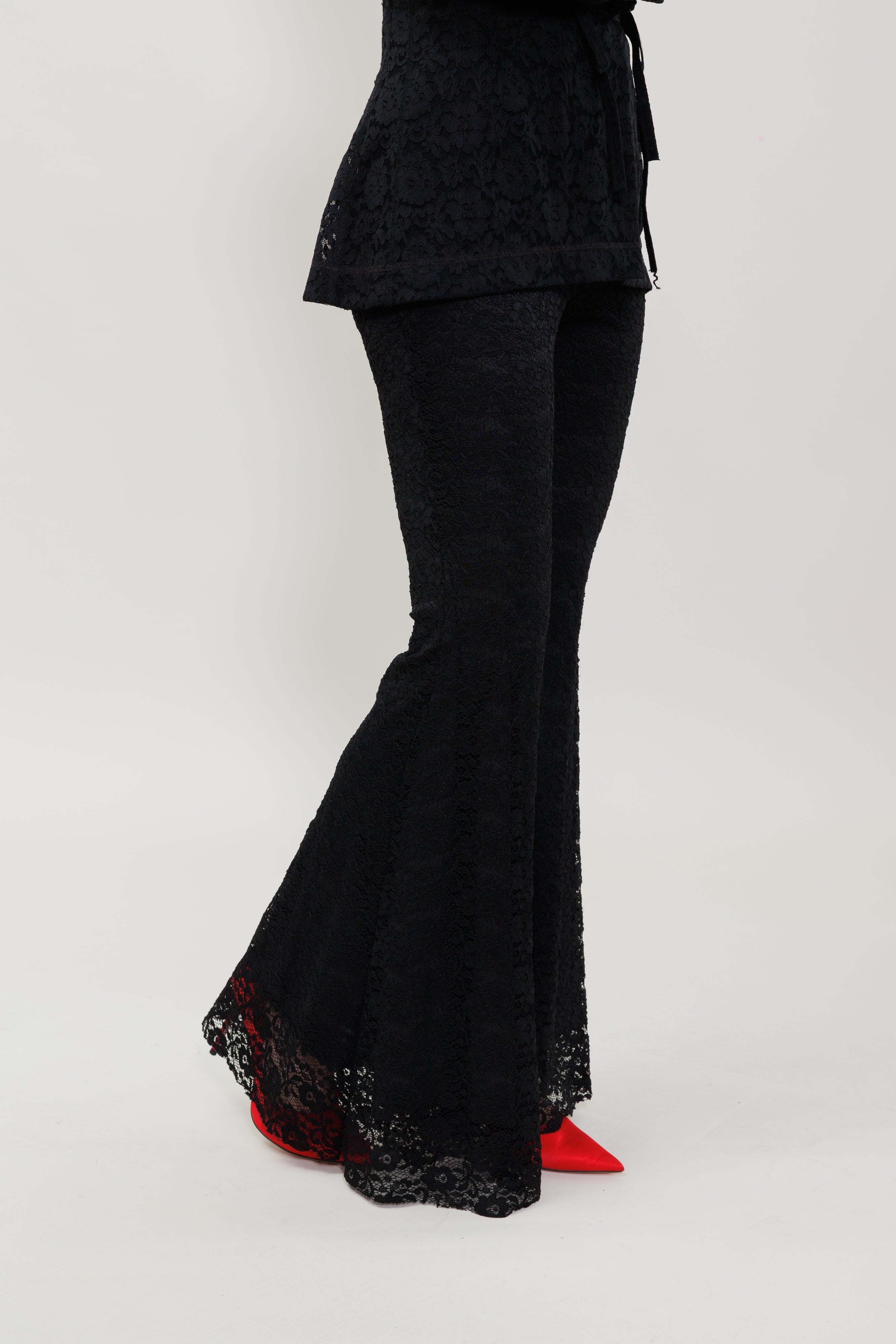 Black lace flared pants with red lining and red pointed shoes, shown on a model from the waist down against a light gray studio background.