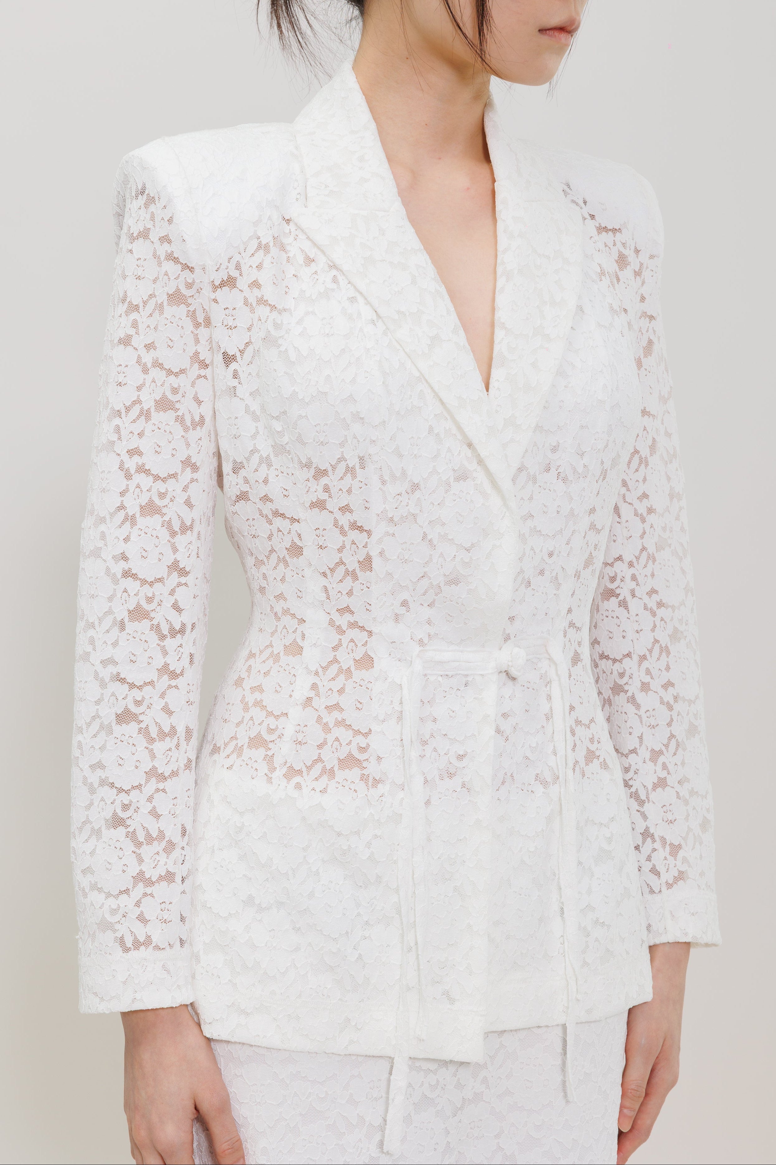 A woman wearing a white lace long-sleeve wrap blazer with a matching tie belt, shown from the chest down against a plain light gray studio background.
