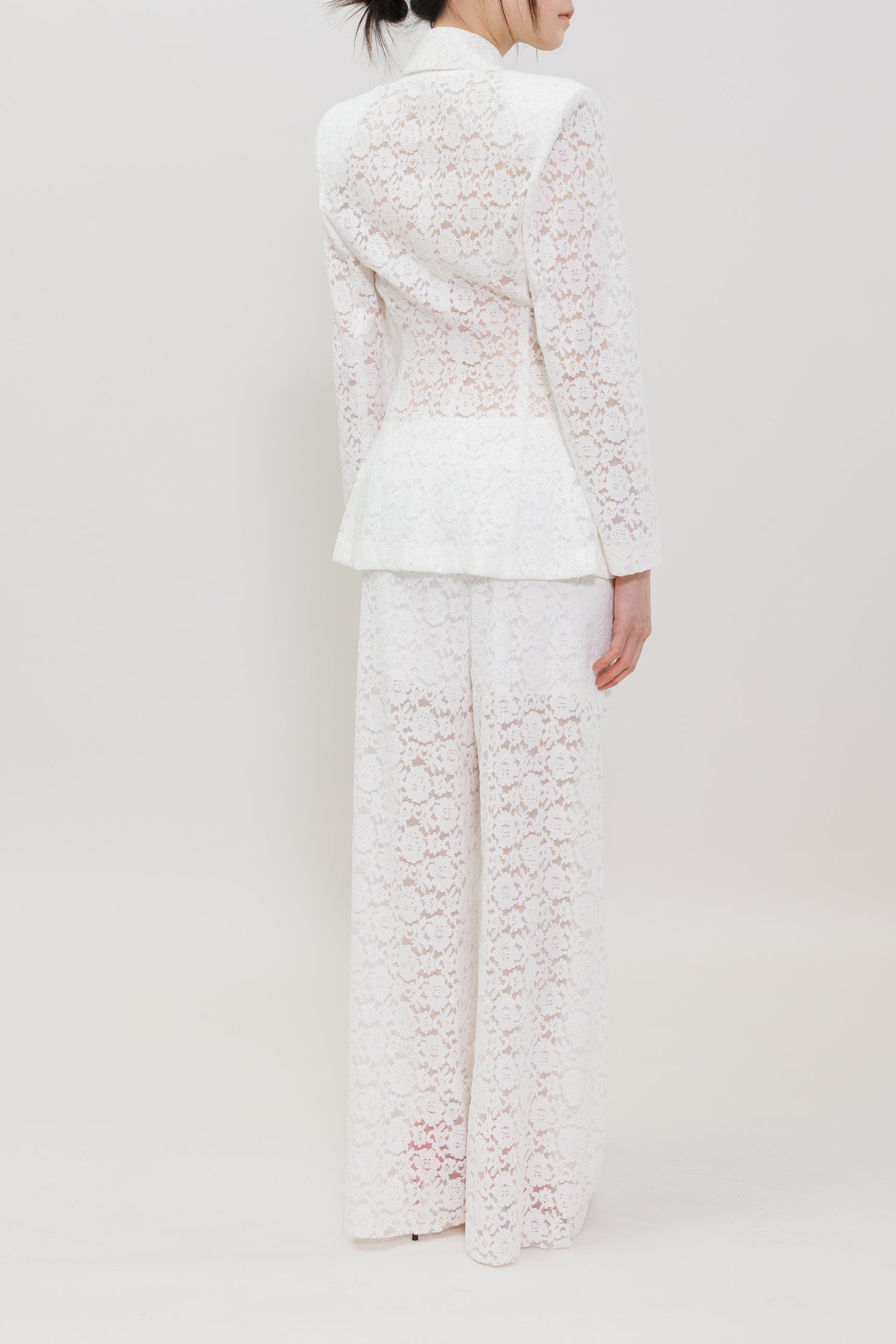 A model viewed from the back wearing a white lace suit with a structured blazer and wide-leg trousers, set against a plain light gray studio background.