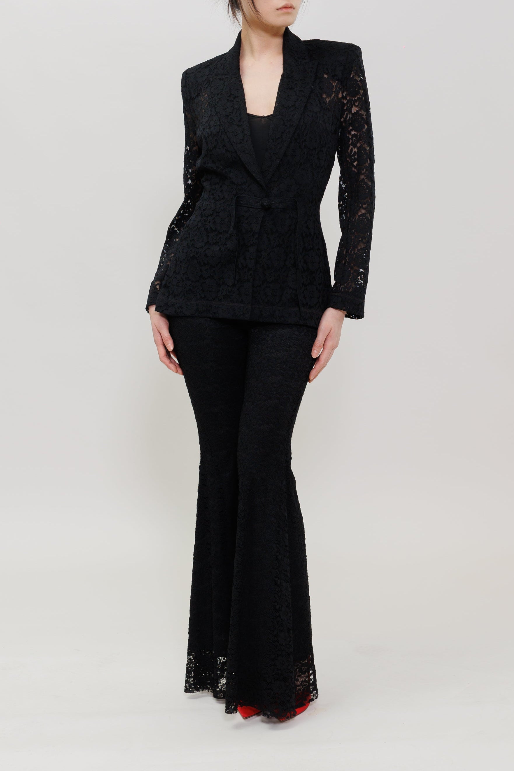 A woman modeling a black lace blazer and matching flared pants against a plain light gray studio background.