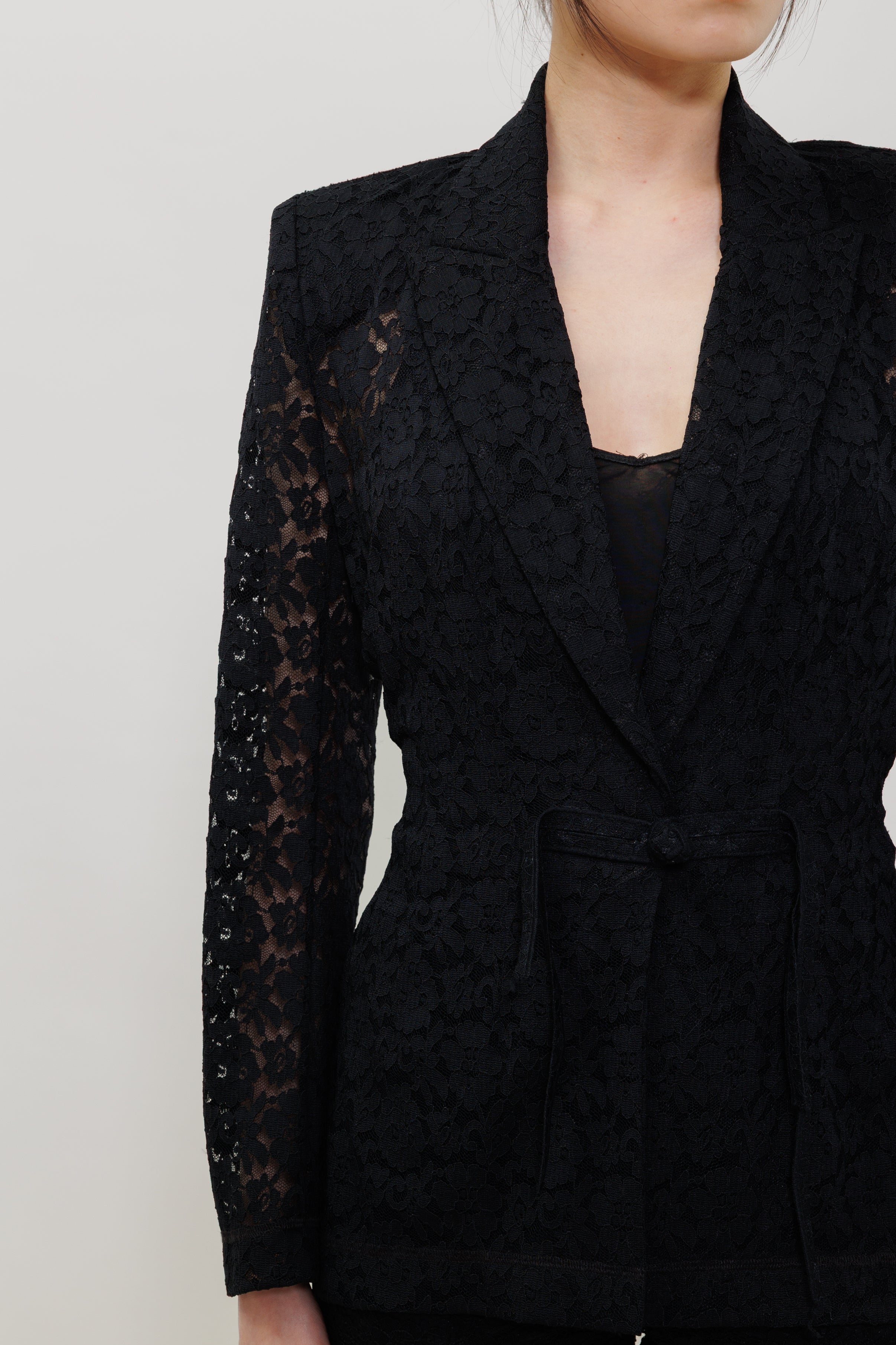 A close-up of a model wearing a black lace blazer with structured shoulders, sheer lace sleeves, and a matching waist tie, set against a plain light gray background.