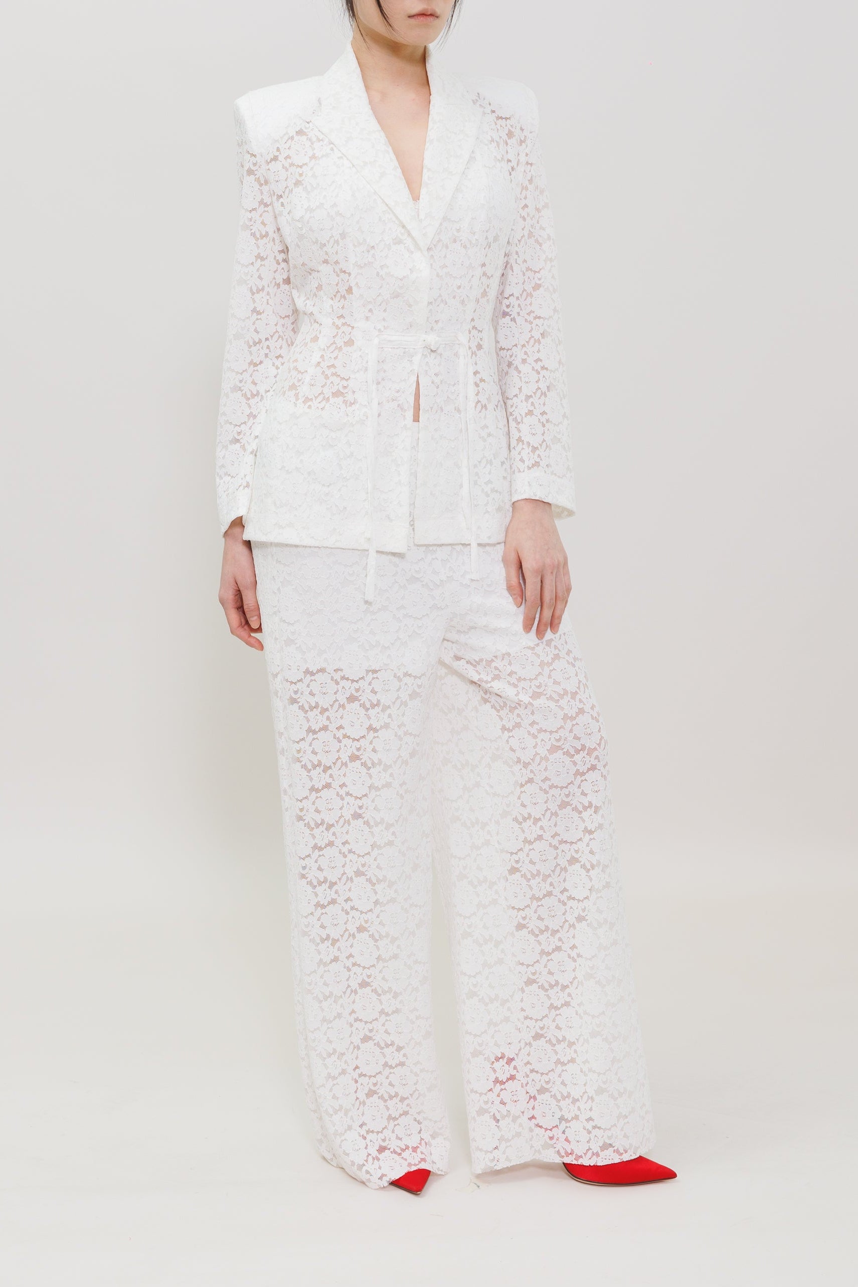 A model stands against a plain white studio background wearing a white lace suit consisting of a blazer and wide-leg trousers, paired with bright red pointed shoes.