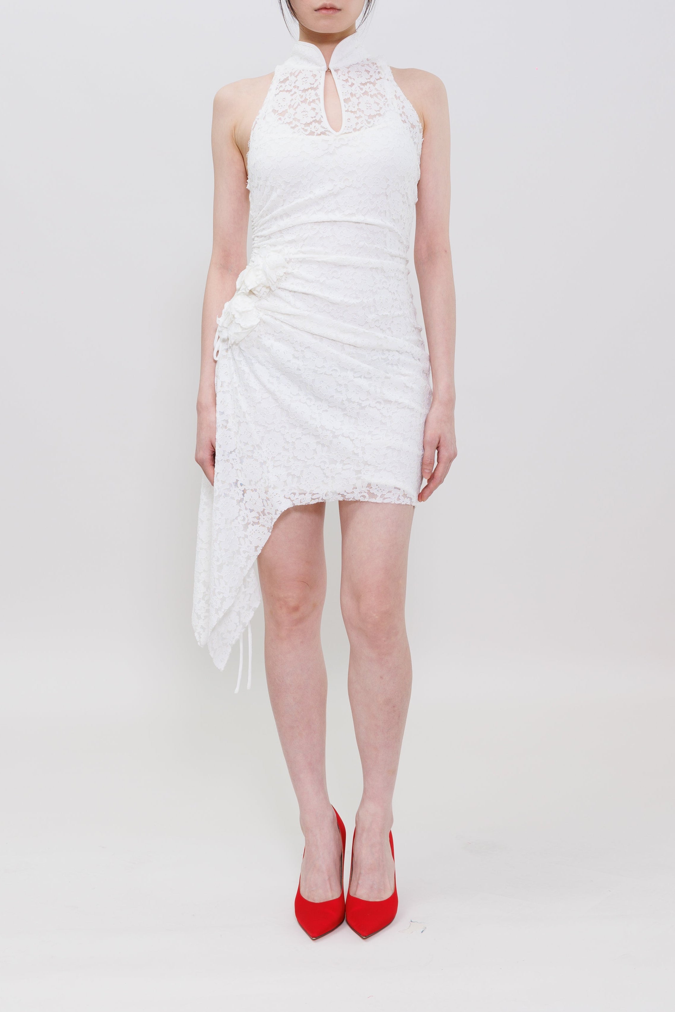 A model stands against a seamless white studio background wearing a white sleeveless lace dress with a high mock neck and red pointed-toe heels.