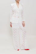 A model stands against a plain white studio background wearing a white lace suit consisting of a blazer and wide-leg trousers, paired with bright red pointed shoes.