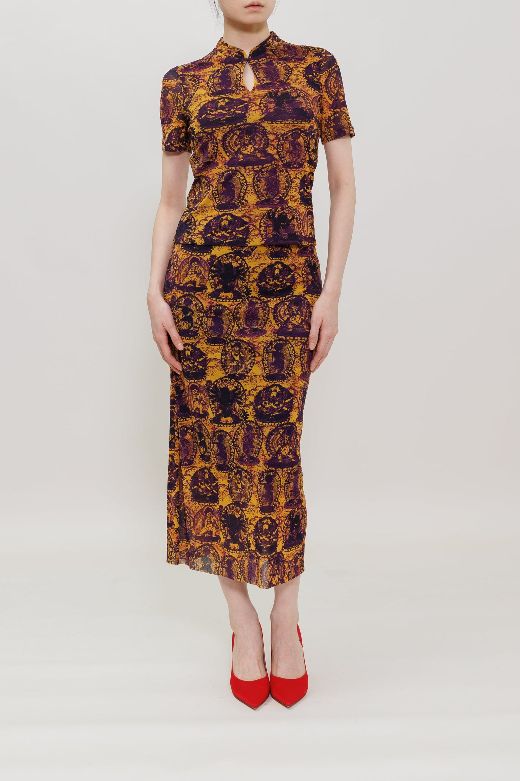 A model wearing a short-sleeved midi-length cheongsam dress with a yellow and dark purple baroque pattern, paired with red pointed-toe heels, set against a plain light gray studio background.