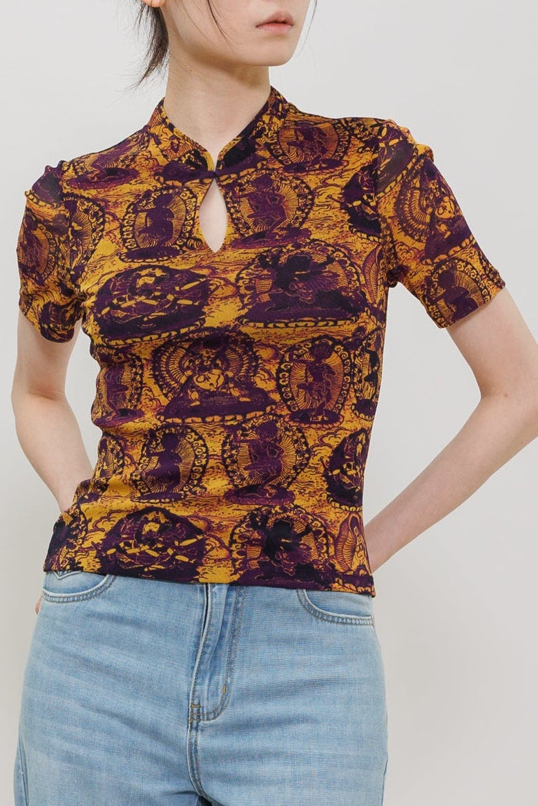A model wearing a short-sleeve, high-neck top with a purple and gold baroque pattern, paired with light-wash wide-leg jeans, shot against a plain light gray studio background.