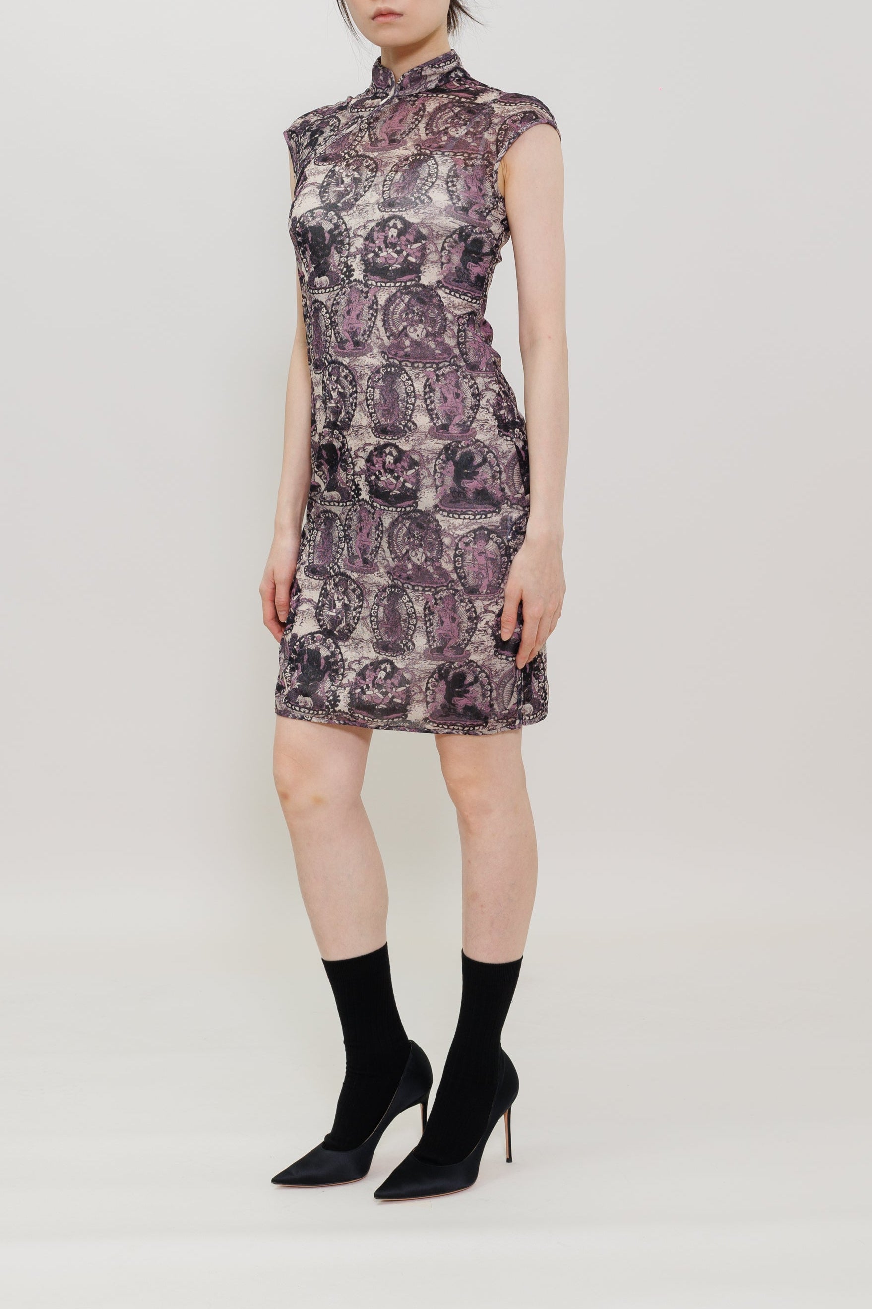 A model stands against a plain light gray studio background wearing a sleeveless high-neck mini dress with a purple and gray abstract pattern, paired with black ankle socks and black pointed heels.