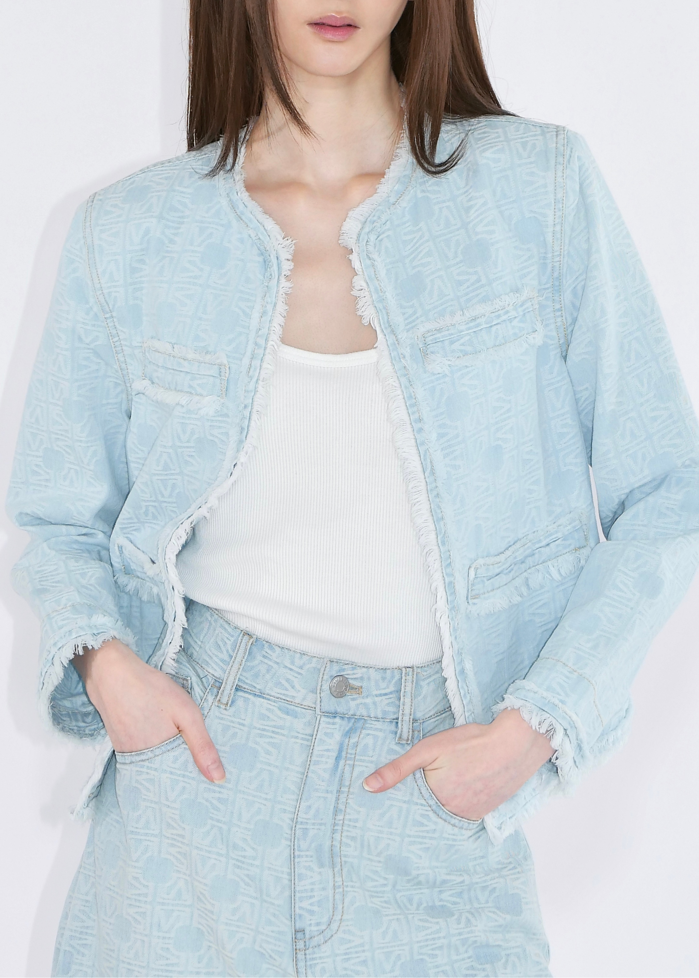 A woman wearing a light blue denim jacket with raw frayed edges over a white tank top and matching light blue denim jeans, photographed against a plain white studio background.