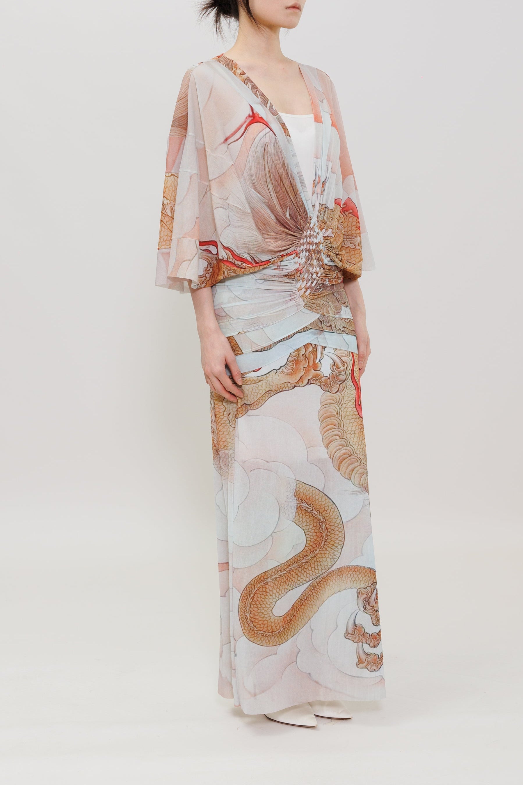 A woman stands in profile wearing a long, loose-fitting dress with a soft dragon print in beige, white, muted orange, and light blue on a plain light gray studio background.