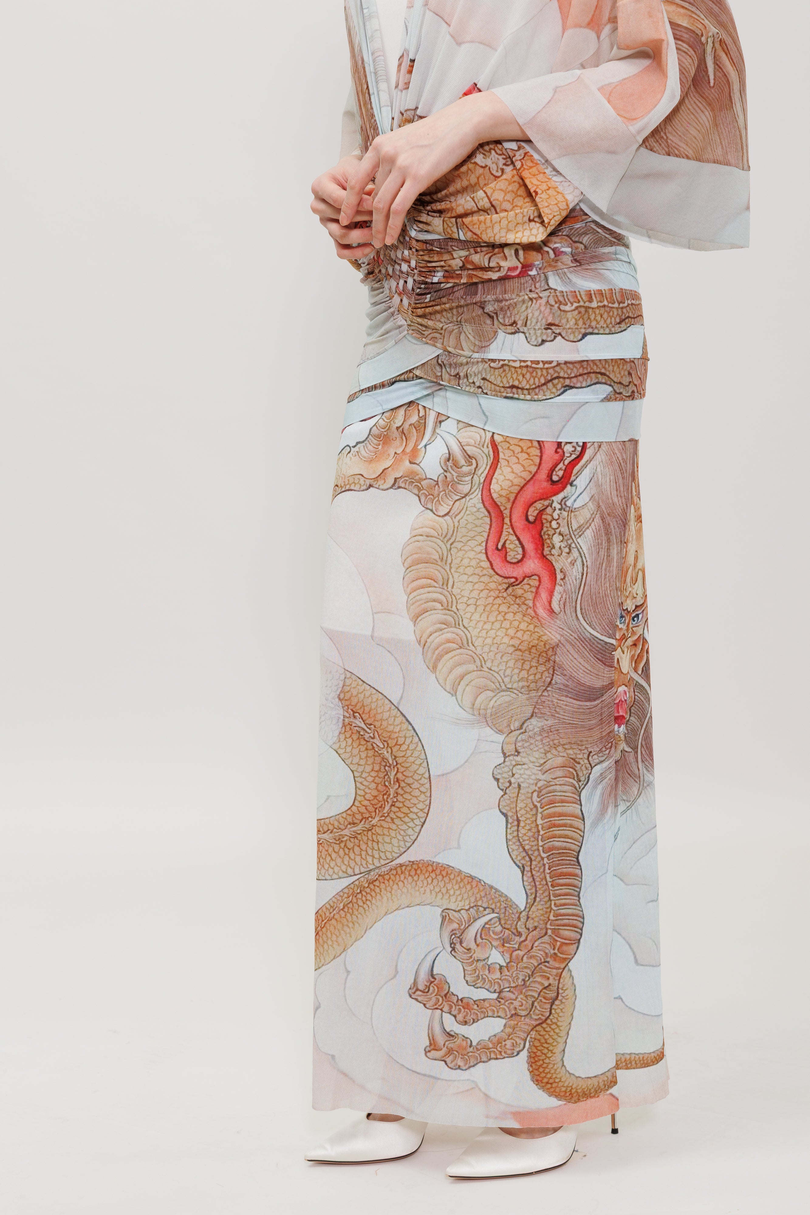 A model wearing a long, sheer dress with a detailed dragon print in gold, brown, and red on a white background, paired with white pointed shoes against a light gray studio backdrop.