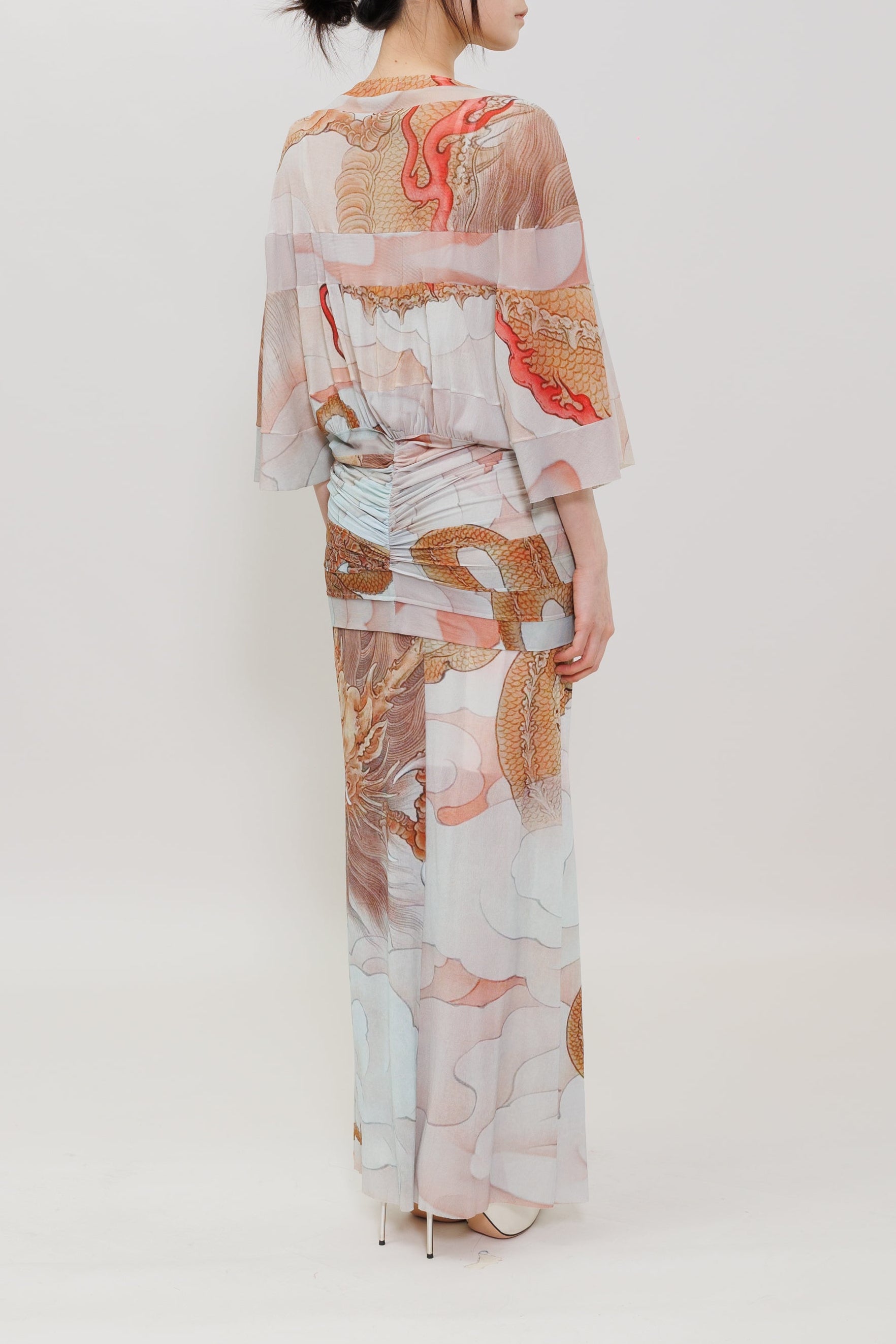 A woman stands with her back to the camera wearing a long, flowing kimono-style dress with wide sleeves and an dragon print in soft pink, beige, white, and rust tones against a plain light gray studio background.