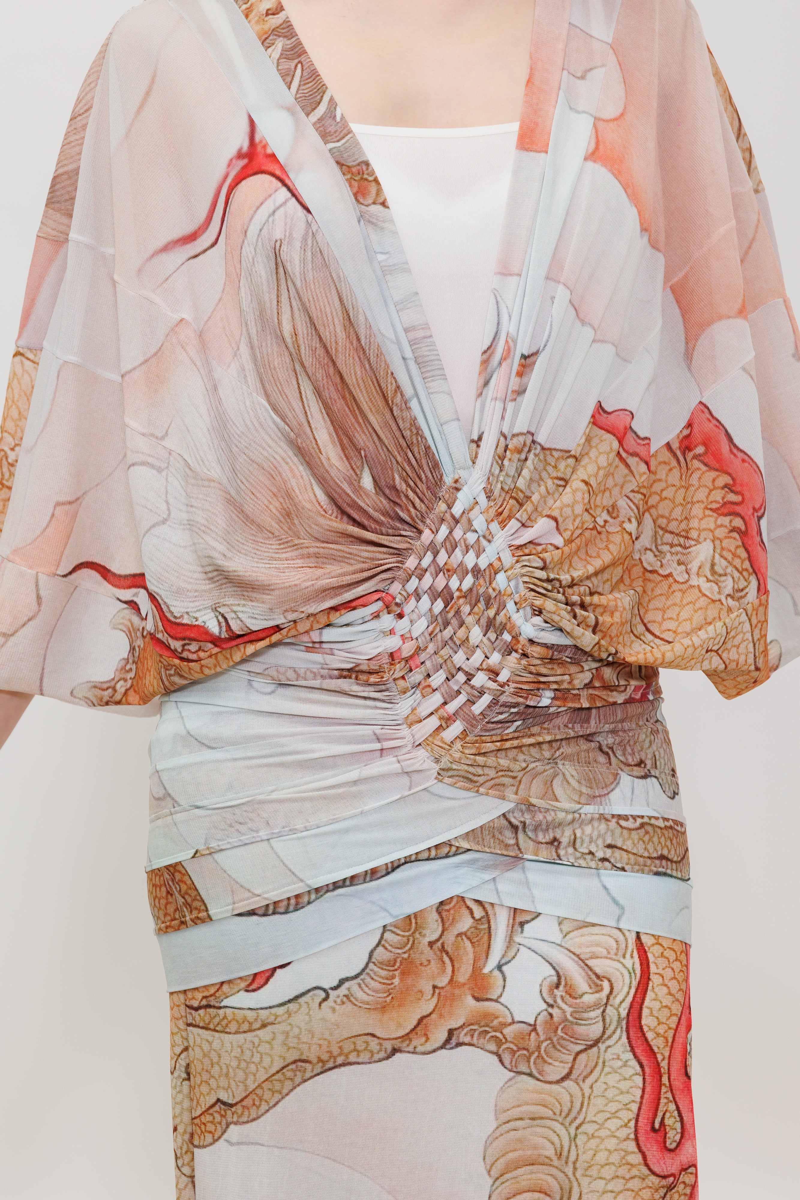 A close-up of a model wearing a sheer, wrap-style garment with a dragon print in beige, brown, and red, tied at the waist over a white top.