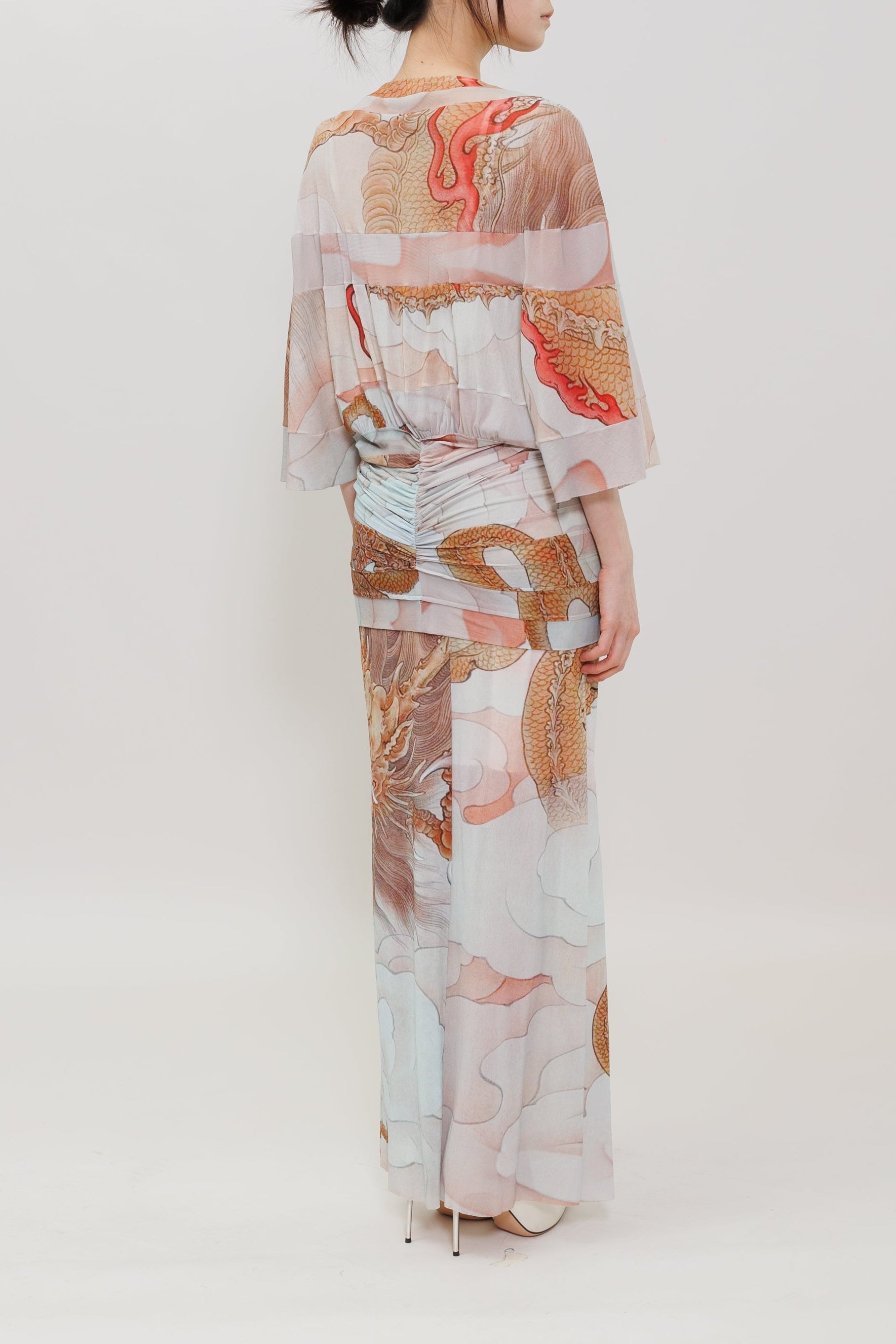 A woman stands with her back to the camera wearing a long, flowing kimono-style dress with wide sleeves and an abstract print in soft pink, beige, white, and rust tones against a plain light gray studio background.