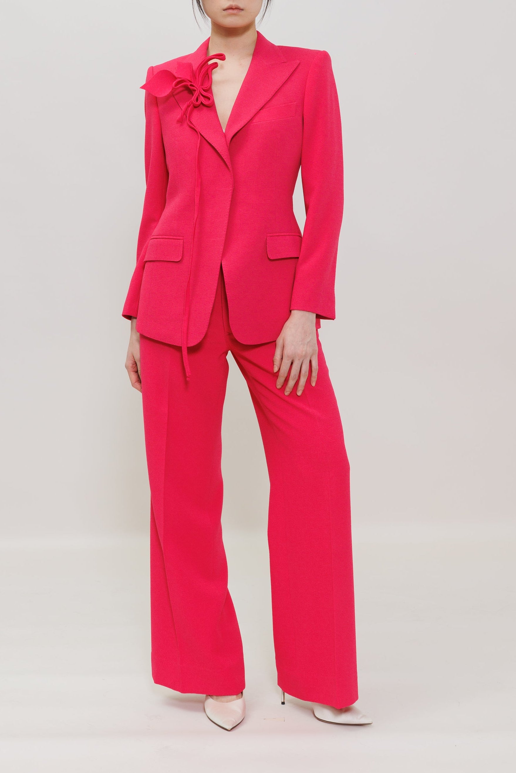A model stands against a light gray studio background wearing a bright pink tailored pantsuit with a structured blazer featuring a large ruffle detail on one shoulder and matching wide-leg trousers.