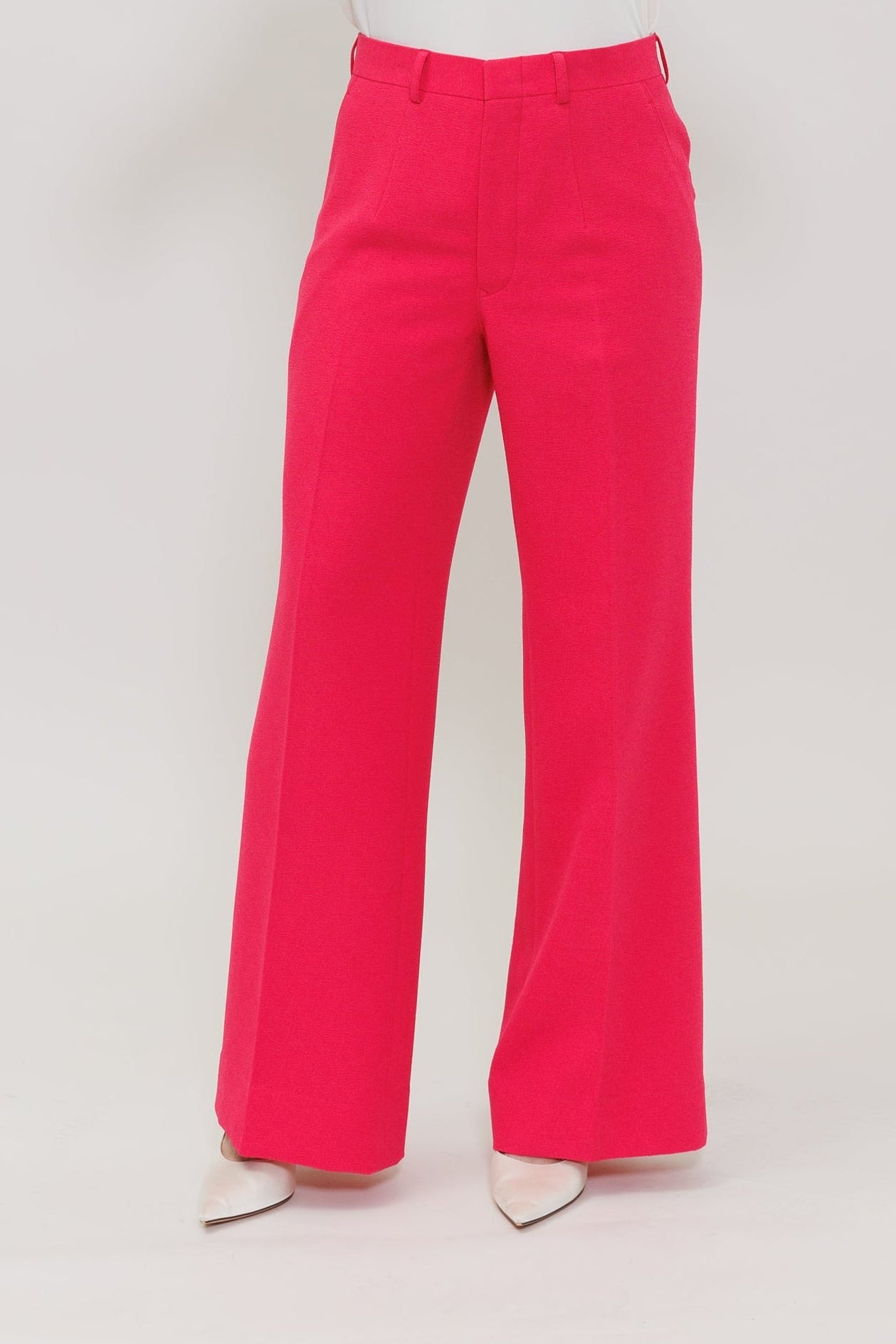 A model wearing bright pink wide-leg trousers and a matching pink blazer, holding up a white undergarment to show the waistband and front of the pants against a light gray studio background.
